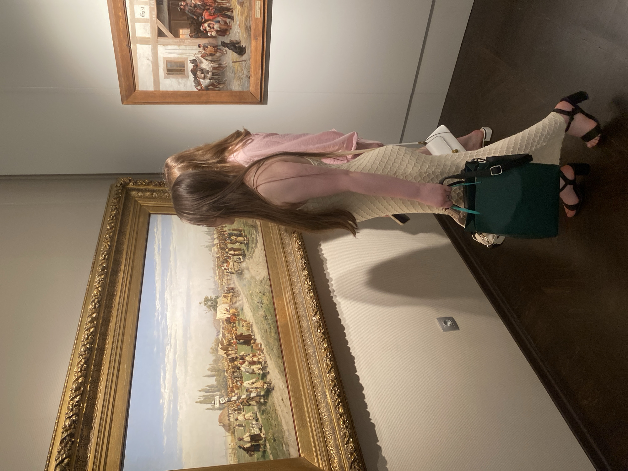 A woman and a child are standing in front of two paintings at a museum. The paintings are both framed with ornate gold frames. The woman is wearing a cream-colored dress and black sandals and holding a green bag. The child is wearing a pink shirt. The museum has light gray walls and a dark wood floor. A square light switch is visible on the wall. Based on visual elements and potential gallery affiliations, this could be the Yekaterinburg Museum of Fine Arts, Russia, though confirmation requires image metadata or location-specific knowledge.