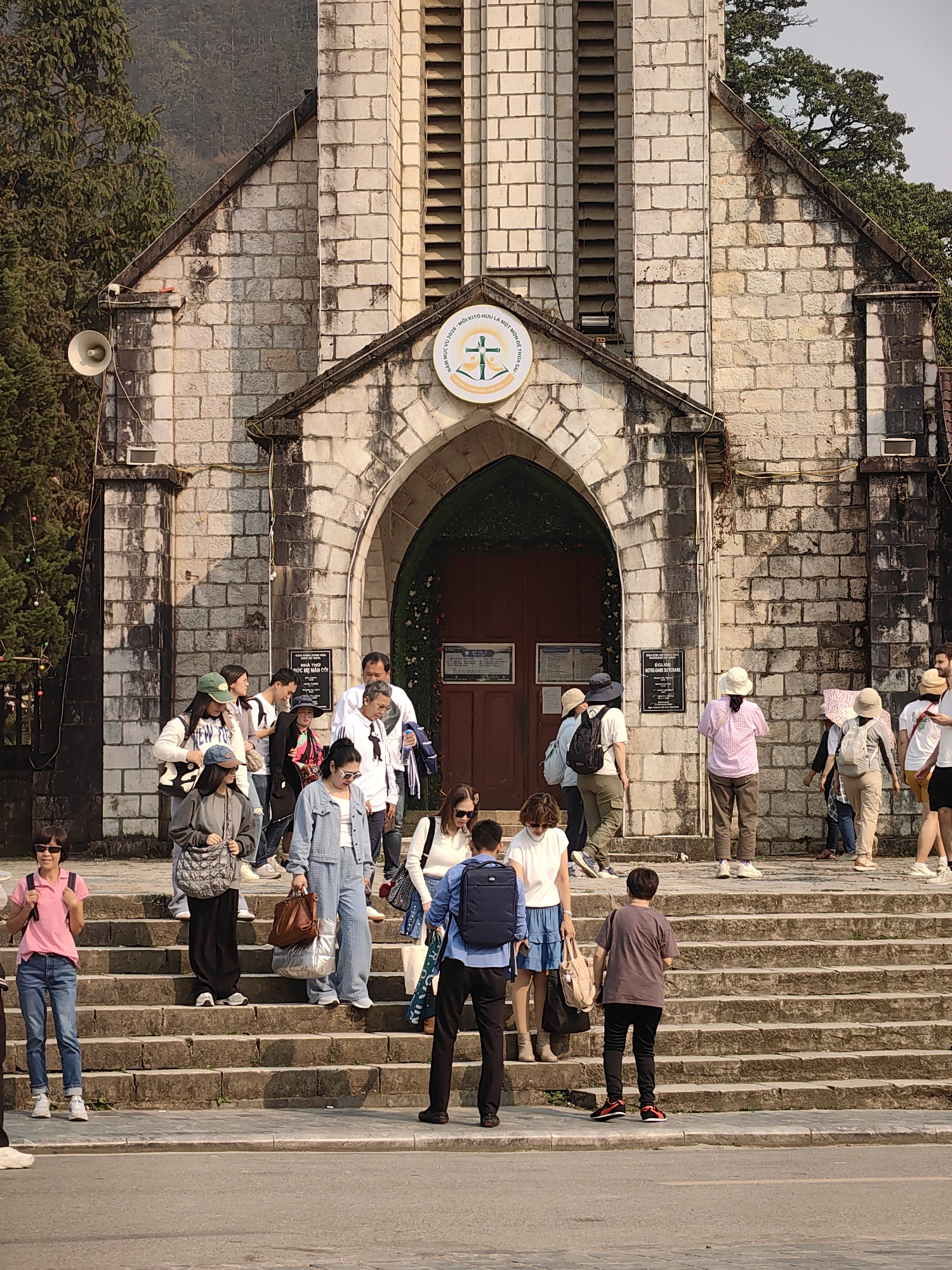 The image displays the Sa Pa Stone Church (Holy Rosary Church) in Sa Pa, Vietnam. The structure is built from light-colored, rectangular stone blocks, featuring a prominent central arched entrance and a bell tower section with vertical louvers. Above the arch, a circular emblem with a cross and Vietnamese text, including "NHÀ THỜ ĐỨC MẸ MÂN CÔI" (Our Lady of the Rosary Church), is affixed. Two informational plaques are visible on the wall to the right of the entrance.
A wide, multi-tiered stone staircase, showing signs of wear, leads from the paved street level up to the church entrance. Approximately twenty to twenty-five individuals are present on and around these steps. The majority are adults, dressed in casual clothing, including t-shirts, jeans, shorts, and light jackets. Several individuals carry backpacks, shoulder bags, or purses. People are observed ascending and descending the stairs, with some congregating near the church entrance and others standing on lower steps or at the base. Specific individuals include a woman in a pink polo and blue jeans on the lower left, facing the camera; a man with a dark backpack at the bottom center, facing away; and a person in a dark t-shirt with black pants and red-soled shoes at the bottom right, also facing the steps.
Pine trees and other foliage flank the church, with distant mountains or hills visible in the background under clear daylight conditions. A white horn loudspeaker is mounted on the left exterior wall of the church, and some decorative string lights are visible on the adjacent tree. The overall scene depicts a public gathering point, likely a tourist attraction.