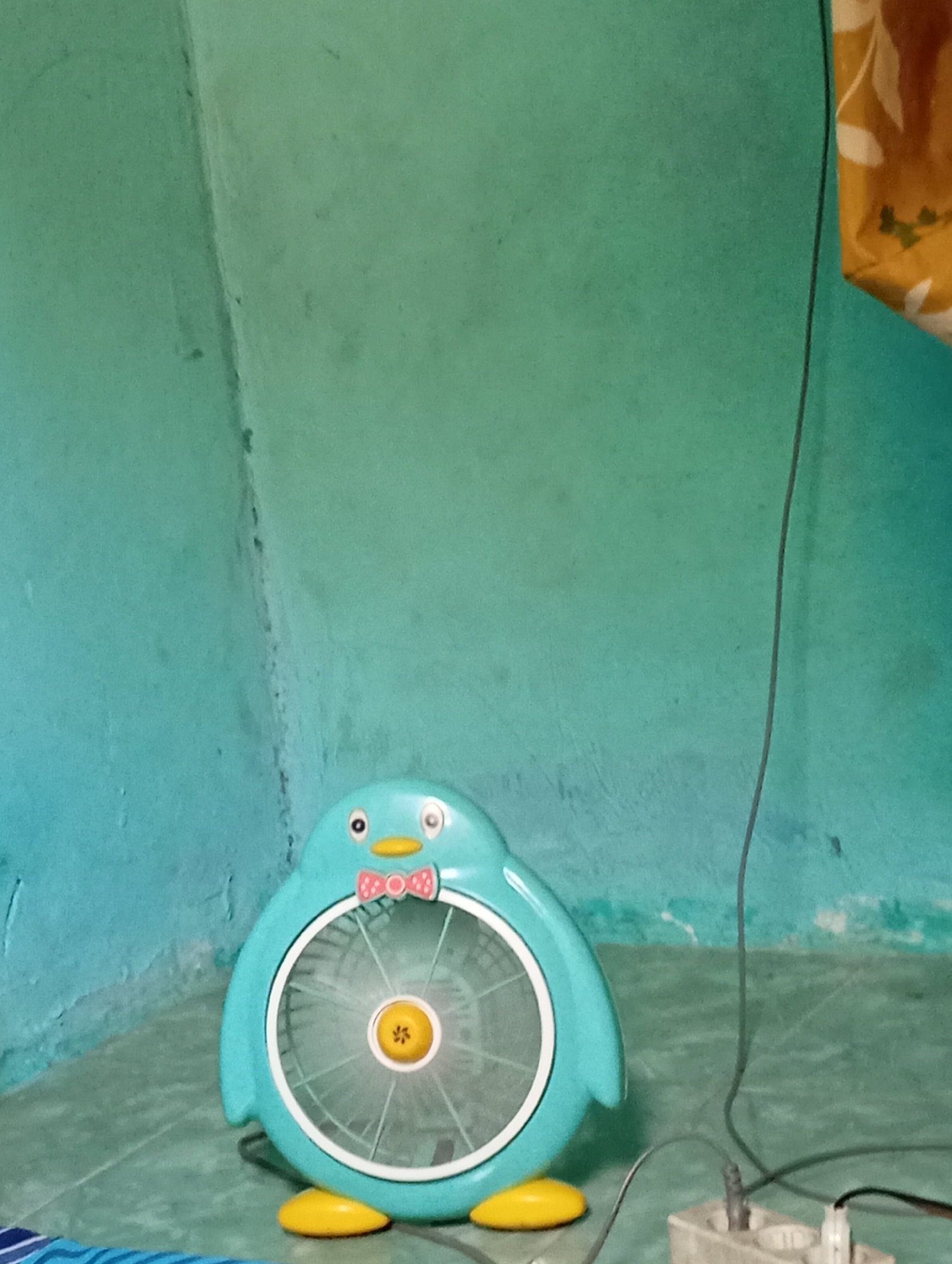 A turquoise penguin-shaped fan is positioned on a tiled floor against a teal-colored wall. The fan has yellow feet, a pink bowtie, and orange eyes. A dark electrical cord runs from the fan to a white power strip in the lower right corner of the frame. A yellow and orange patterned fabric is draped over the top right edge of the image. The environment appears to be an indoor domestic setting.