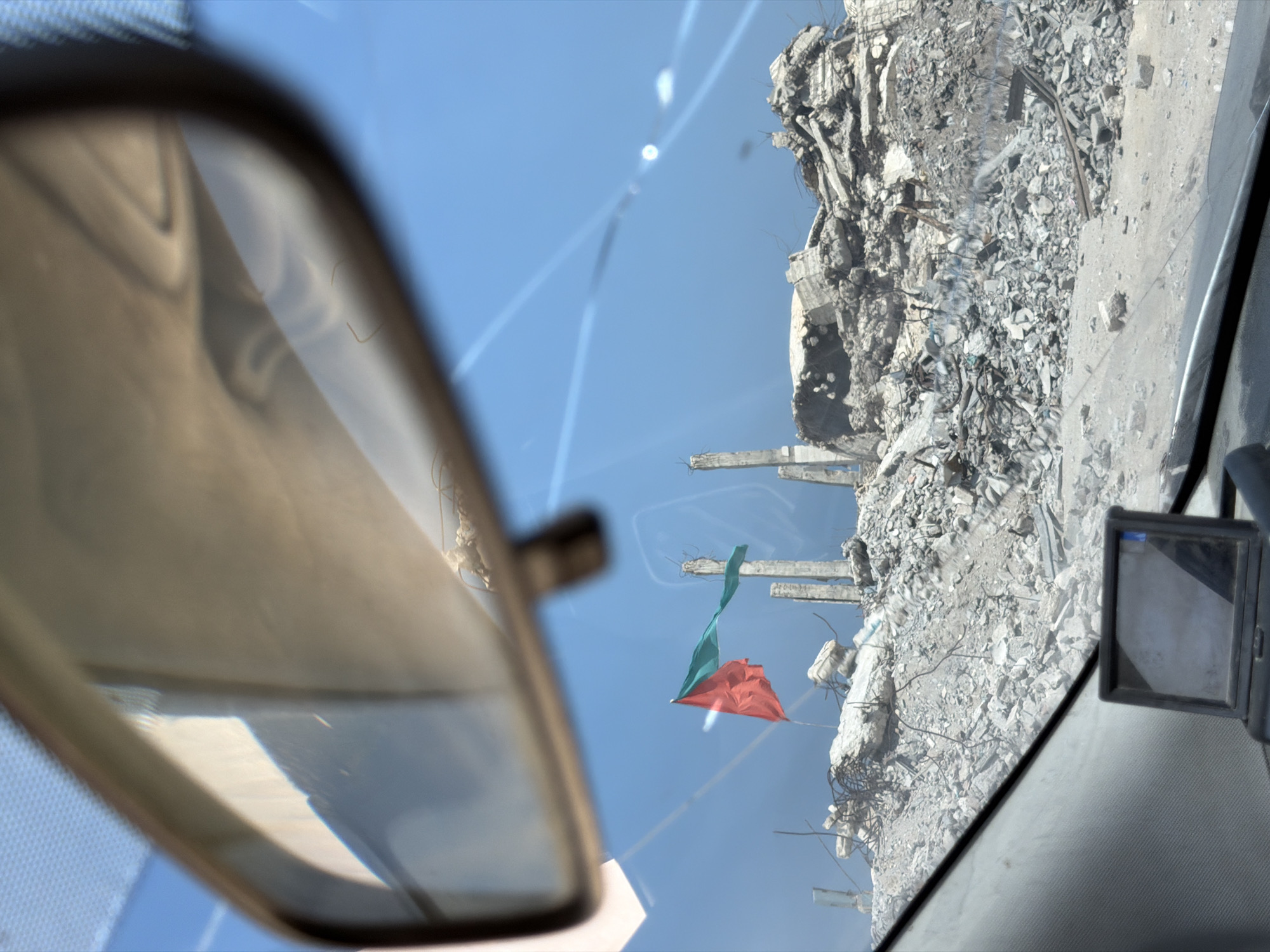 The image depicts the aftermath of destruction in the city of Jabalia, Palestinian Territories. In the foreground, the dashboard of a vehicle is visible, with a small black electronic device, possibly a GPS or dashcam, mounted on it. A rearview mirror is prominently positioned, reflecting a hazy, light-colored interior and the bright blue sky.
Beyond the vehicle's windshield, the scene unfolds as a landscape of extensive destruction. Piles of rubble, concrete debris, and twisted metal rebar dominate the terrain. Several damaged concrete structures, appearing to be the remnants of buildings, stand as broken columns against the sky. A flag, split into two colors, red and green, flies from what seems to be a makeshift flagpole amidst the debris. The environment appears arid and dusty, under a clear, bright blue sky. Sunlight creates lens flares and reflections on the windshield and in the mirror. The overall impression is one of devastation and the stark reality of a war-torn environment.
