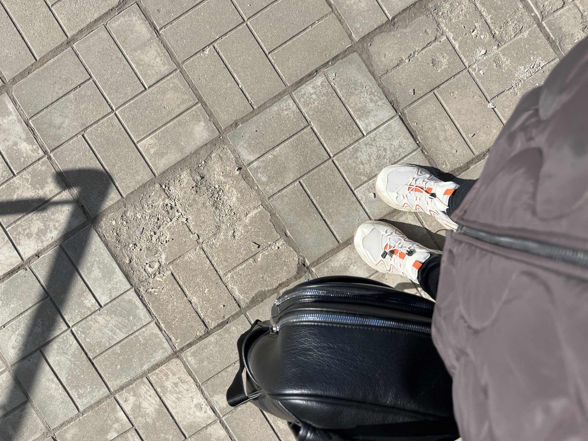 The image shows a portion of a paved pedestrian walkway with interlocking gray concrete bricks, with an area on the left where some of the bricks are missing and replaced by loose dirt and debris. Two feet are visible, wearing white sneakers with orange accents and black pants. Adjacent to the feet is a black leather backpack or bag with a prominent zipper. Part of a light-colored, quilted jacket is visible on the right. The scene is illuminated by sunlight, casting a shadow on the left side of the frame. The image does not provide enough information to confirm that this image was taken in the city of Klin, Russia.