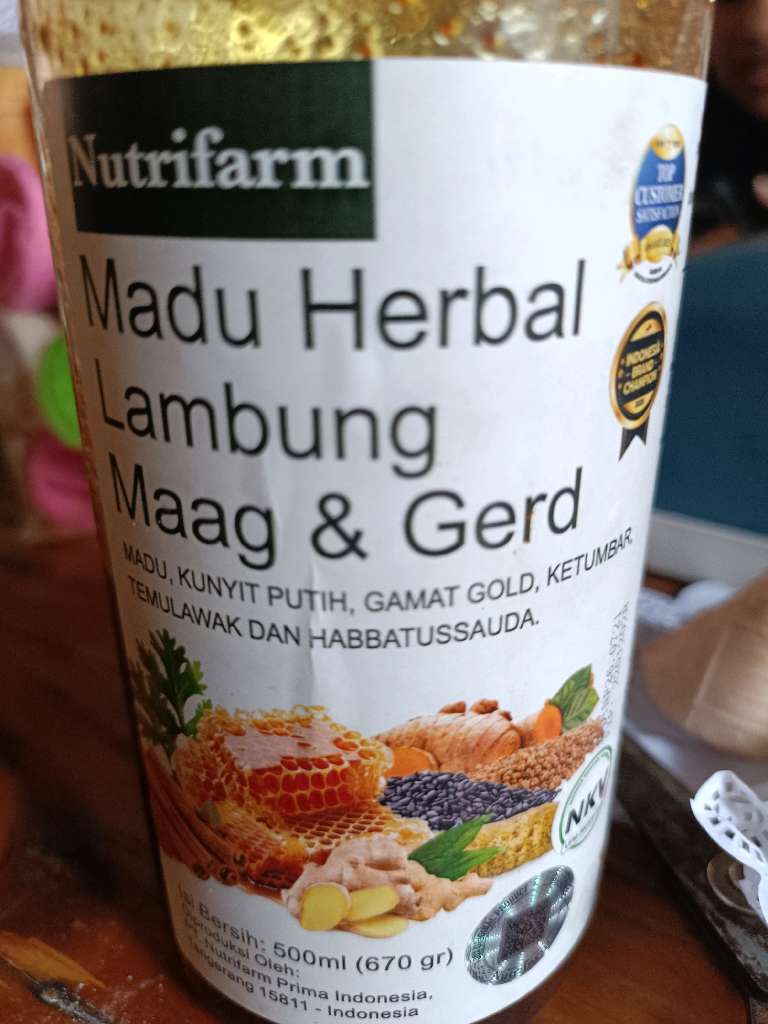 The image shows a close-up of a bottle of "Nutrifarm Madu Herbal Lambung Maag & Gerd" with Indonesian text on the label. The bottle is positioned on a wooden surface, likely a table. The label features an illustration of honeycomb, ginger, and other ingredients, as well as seals indicating "Top Customer Satisfaction" and "Indonesia Brand Champion." There are no visible people or specific environmental details that would indicate a location within Jatitengah, Indonesia.