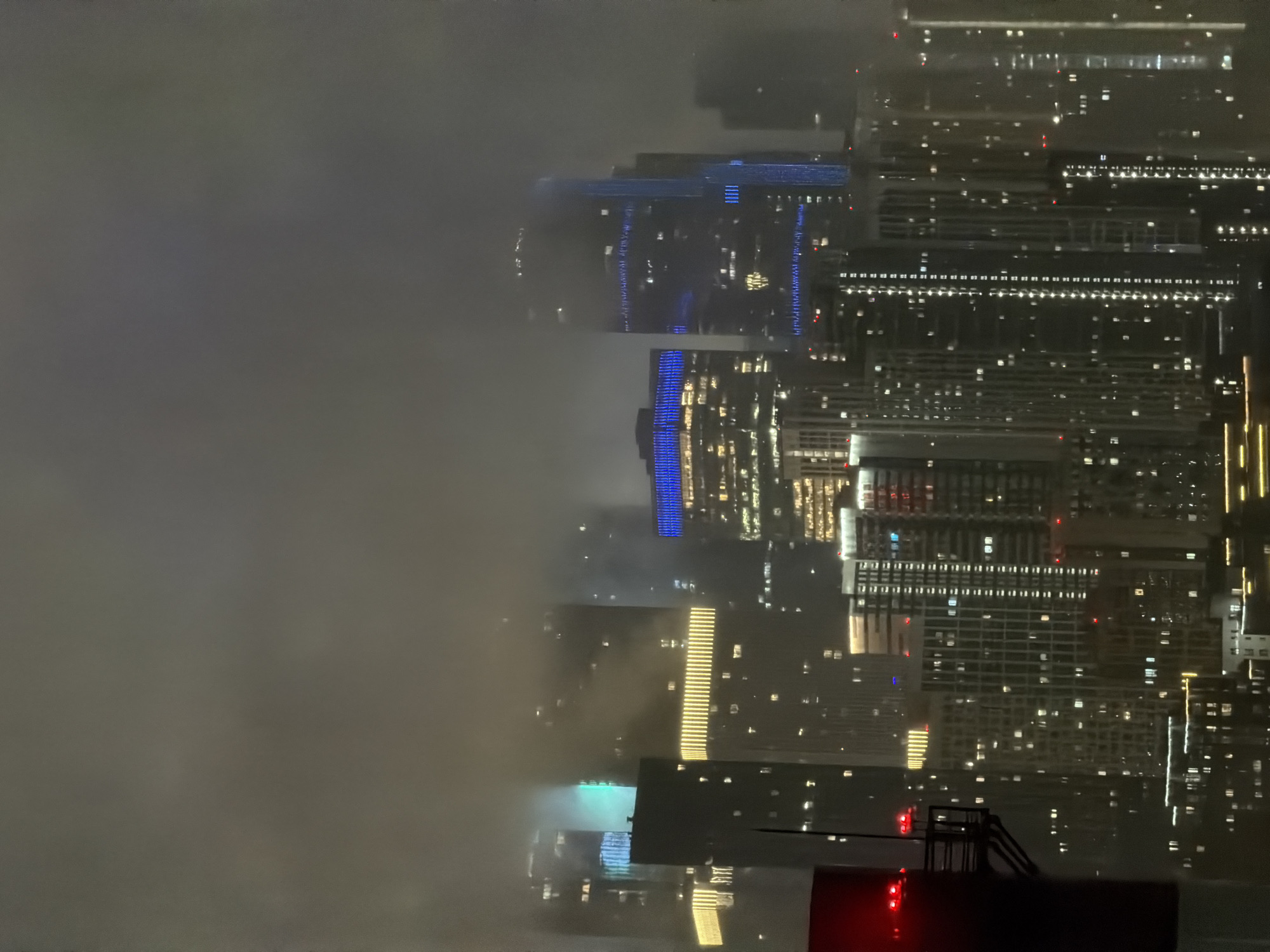 The image depicts a nighttime cityscape featuring numerous high-rise buildings in Khimki, Russia, partially obscured by fog or clouds. The architecture is modern, with many structures featuring illuminated windows and decorative lighting. Some prominent buildings are highlighted with blue and yellow lights, suggesting a commercial or residential district. In the foreground, on the left side, a dark structure with red lights is visible, possibly part of a tower or antenna. The overall atmosphere is somewhat hazy due to the atmospheric conditions. There are no visible people in the image.
