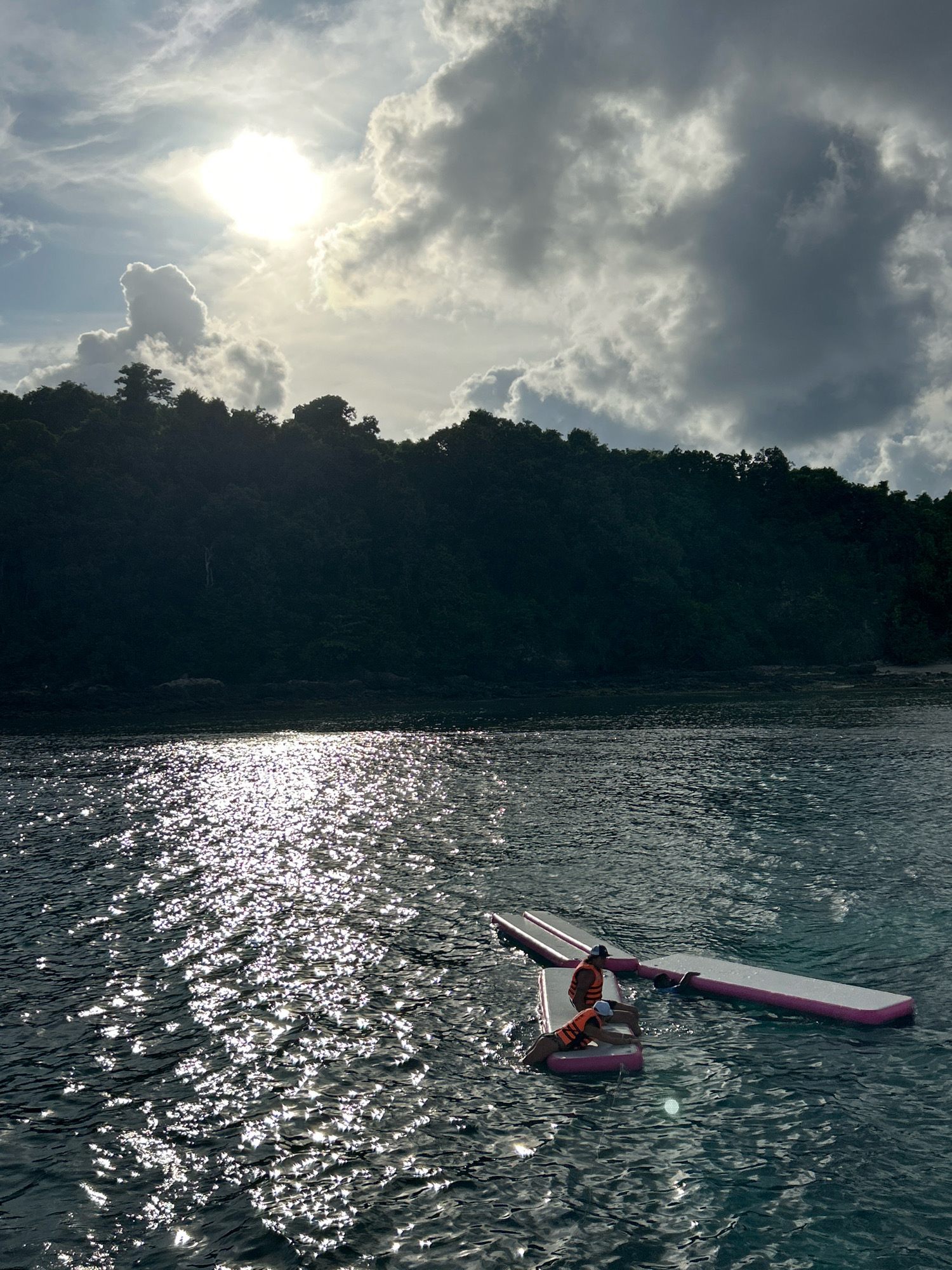 In Rawai, Thailand, a serene ocean scene unfolds under a dramatic sky. The sun, partially obscured by fluffy white and grey clouds, casts a shimmering, dappled light across the dark water. In the foreground, two inflatable water mats, one pink and white, are anchored together. On the left mat, two individuals wearing orange life vests are positioned, one lying prone and the other kneeling. A third individual is visible on the right mat, leaning back. The water is calm, reflecting the light and creating a sparkling effect. Behind the water, a lush green, tree-covered hillside rises, forming a dark silhouette against the bright sky. The coastline is rocky, with some visible rocks at the water's edge. The overall impression is one of tranquil activity on the water, with the natural beauty of the coastline and sky serving as a backdrop.