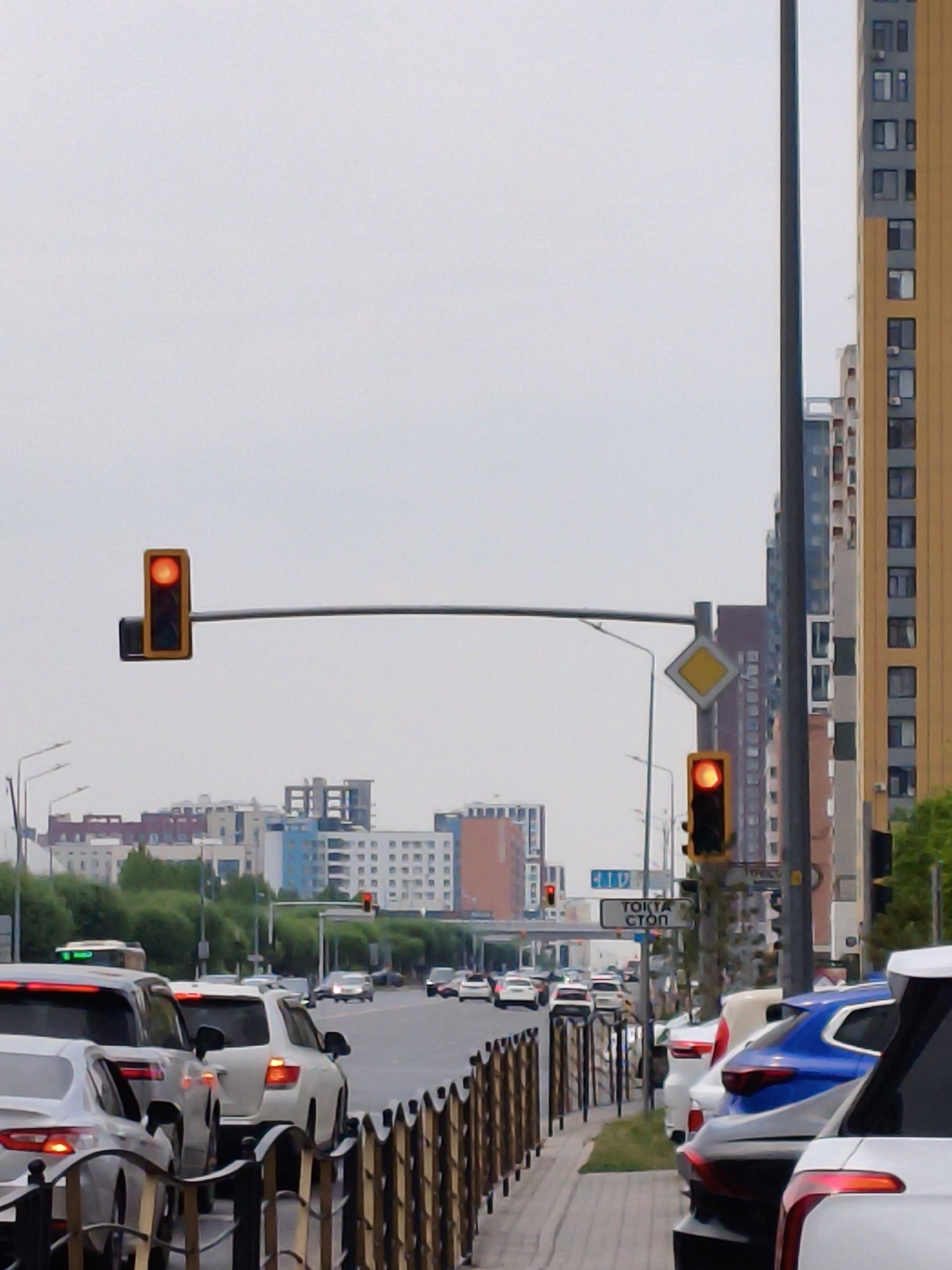 The image depicts a traffic scene in Astana, Kazakhstan, likely during the daytime on an overcast day. In the foreground, a line of cars is stopped at a traffic light. The traffic light is red, indicating that vehicles should halt. The road is lined with modern buildings, some of which are tall and residential, characterized by multiple windows and balconies. The sky is a uniform grey, suggesting cloudy weather. A pedestrian walkway is visible to the right of the road, also bordered by parked cars. A street sign with text in what appears to be Cyrillic script is also present, positioned near a traffic light. The overall impression is one of a busy urban environment experiencing a traffic slowdown.