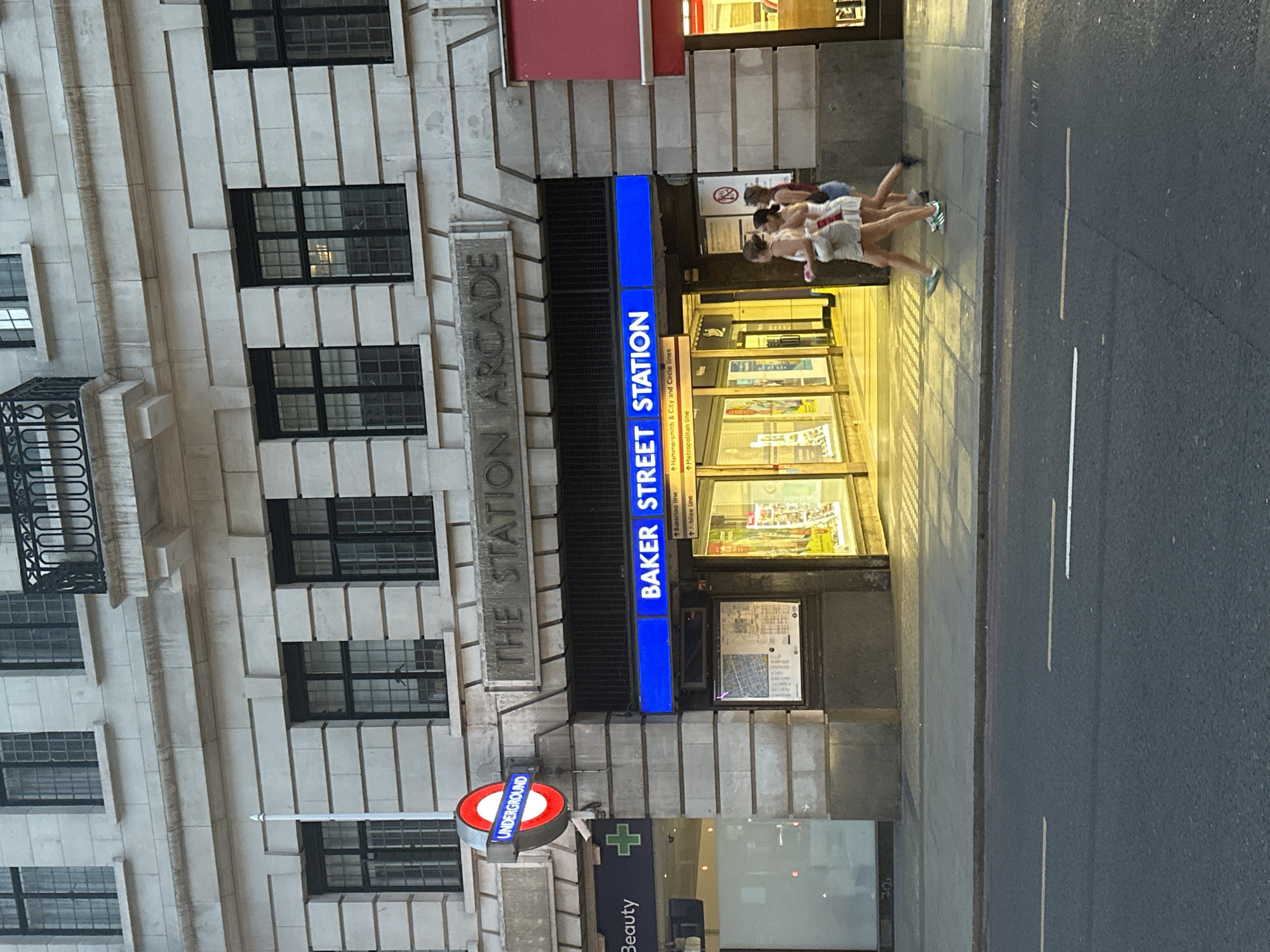 The image shows an entrance to the Baker Street Station in London, United Kingdom. The station is marked by a blue illuminated sign with "BAKER STREET STATION" in white lettering and the round Underground logo sign. The entrance is part of a building with a stone facade and numerous windows. Three people are walking into the station, their bodies blurred due to movement. A pharmacy or beauty store is located next to the station entrance.