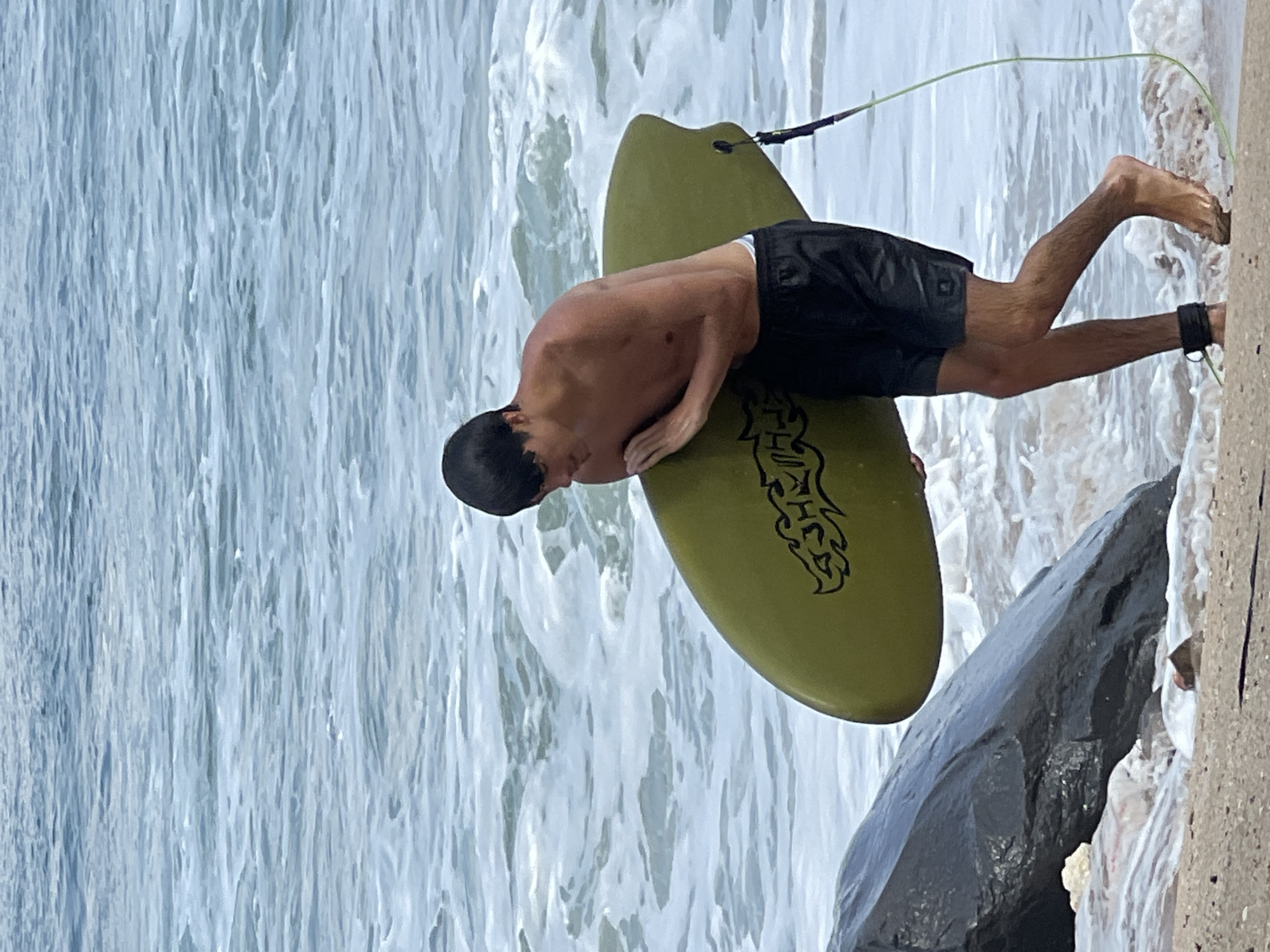 A young man with dark hair and a tan torso is walking on the beach in Kuta, Indonesia, carrying a lime green surfboard. He is wearing black swim trunks and has a black ankle leash attached to his right leg, which is connected to the surfboard. The surfboard has a black graphic design on it. He is walking towards the ocean, which is a pale blue with white foam from the waves. To his left, there is a large dark rock. The sand on the beach is a light tan color.