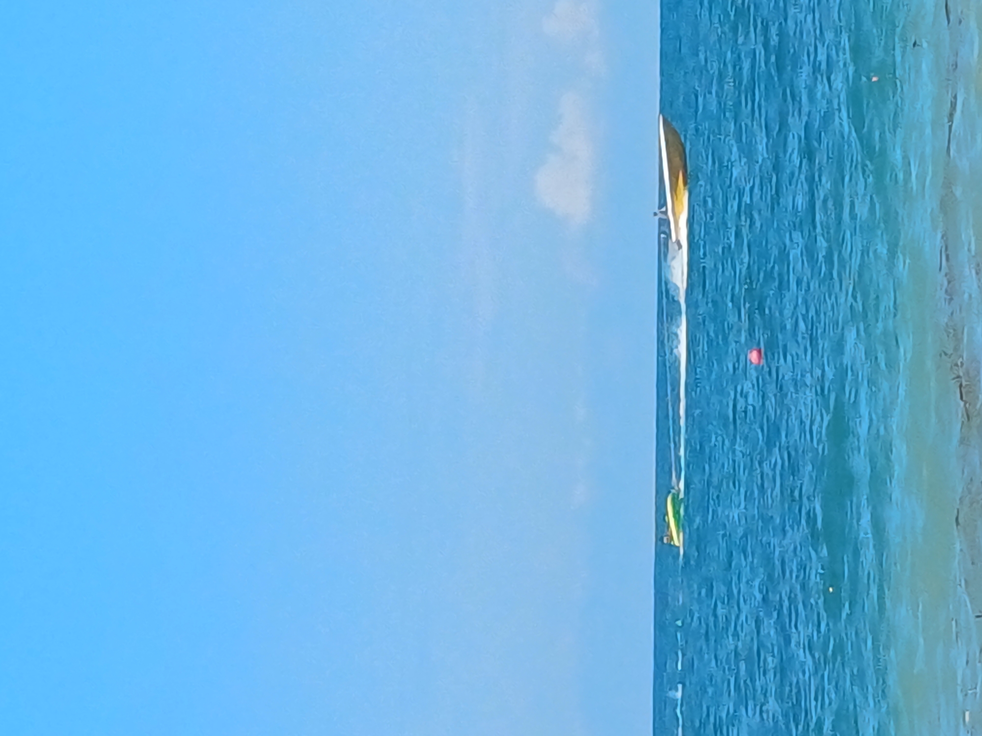 This image captures a bright, sunny day at the sea, likely in Sochi, Russia, given the context provided. The scene is dominated by a vast expanse of clear, blue sky, with a few wispy white clouds scattered across the upper portion. Below the sky lies the tranquil, turquoise sea, with gentle waves rippling across its surface.
In the mid-ground, two distinct watercraft are in motion. To the left, a yellow and green inflatable tube, likely being towed, is being pulled by an unseen vessel, leaving a white wake behind it. Further to the right, a yellow speed boat with a person at the helm is cruising at speed, also generating a significant white wake. The boat appears to be a sleek, modern design, cutting through the water.
In the foreground, the shallow water near the shore is visible, with a hint of the seabed and some scattered debris, possibly seaweed, suggesting proximity to a beach. A small, red buoy is also visible floating on the water between the two main watercraft, indicating a marked area or a navigational point. The overall impression is one of leisure and outdoor recreation on a beautiful day by the sea.