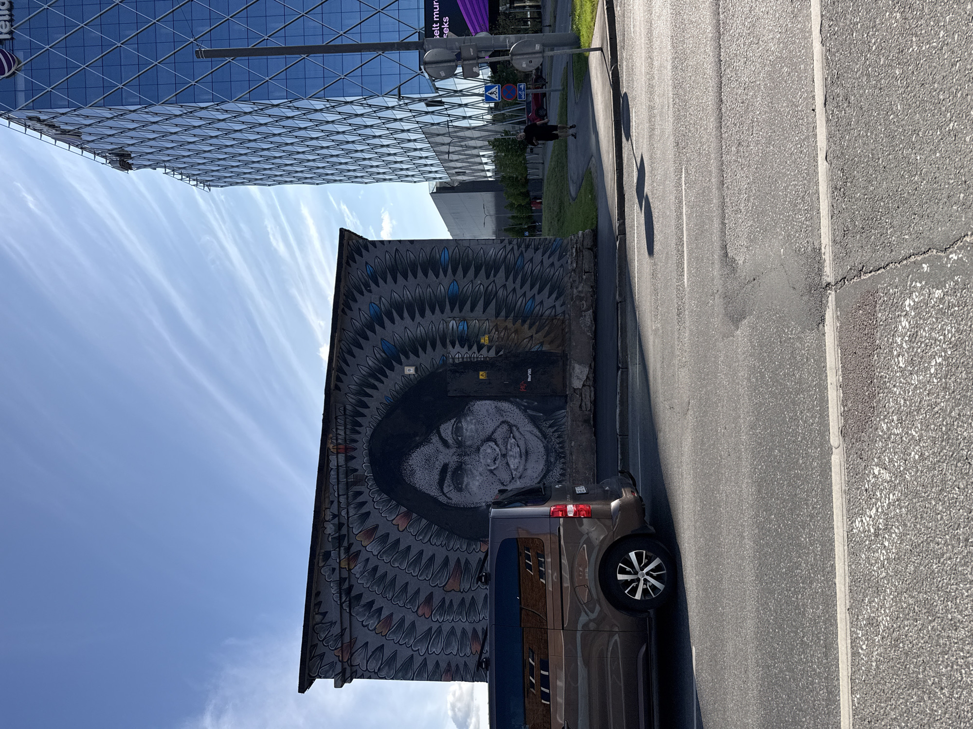 A mural of a woman's face is painted on the side of a grey, weathered building in Tallinn, Estonia. The mural is large and covers most of the building's facade. The woman in the mural has dark hair and is wearing a headdress that resembles feathers. In front of the mural, a brown van is parked, facing away from the camera. To the right of the mural, a modern glass skyscraper rises into the blue sky, reflecting the clouds. A man is jogging on a sidewalk in front of the skyscraper. The street in front of the building is paved with asphalt, and there are white lines indicating lanes and a pedestrian crossing. The sun is casting shadows on the street.