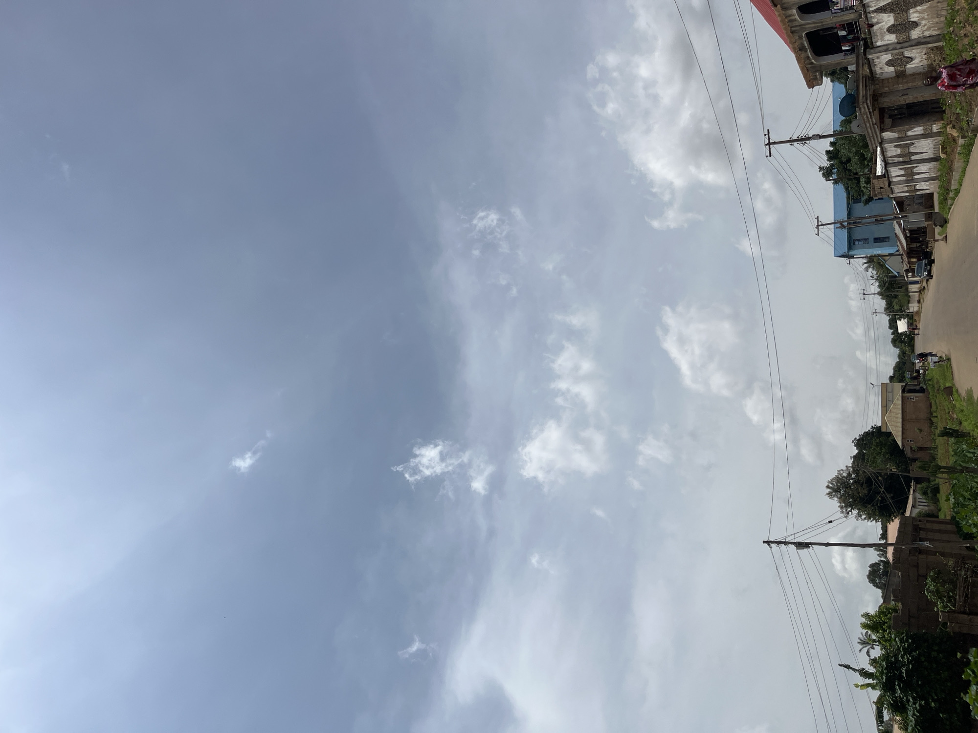 The image depicts a street scene in Bukuru, Nigeria, under a cloudy sky. The road is paved and runs through a built-up area with various structures lining its sides. On the left, there are buildings partially obscured by lush green foliage, including trees and bushes. A prominent wooden pole with electrical wires stretches across the frame, indicating the presence of utility infrastructure. Further down the street, a few cars are visible, suggesting ongoing traffic.
On the right side of the road, several buildings are more clearly visible. One building has a blue facade and a flat roof, while another is constructed with brick and features a red-tiled roof and arched openings. A wall with an ornate design separates the properties. A few individuals are present; one person in a red and white patterned garment is walking on the right side of the road, and another person is visible in the distance on the left. The overall impression is that of a typical urban environment in Nigeria, with a mix of residential and commercial buildings, and evidence of daily life.