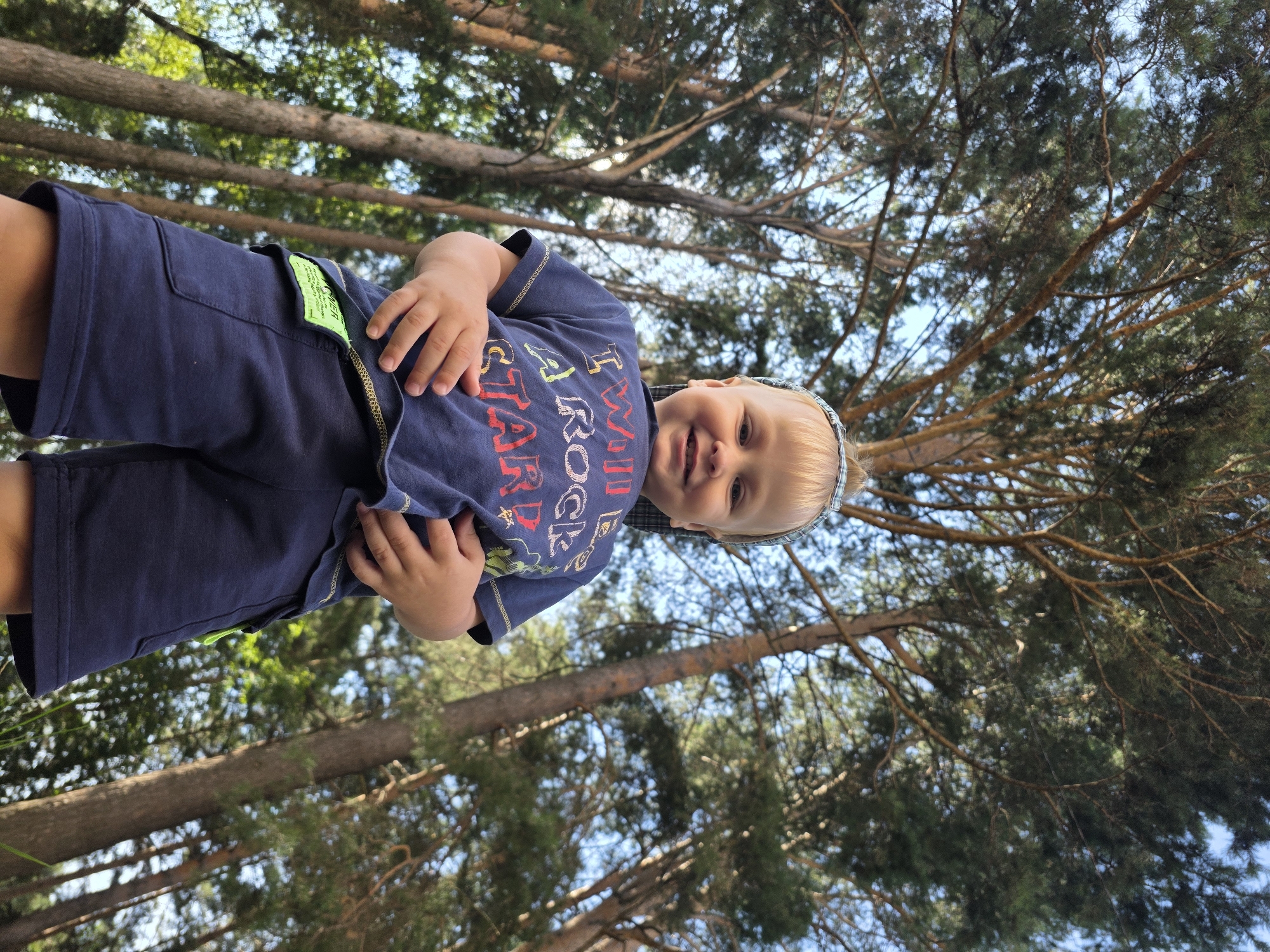A low-angle shot shows a young, blond boy in a navy blue t-shirt and shorts. He is standing outdoors, looking up and smiling at the camera. The t-shirt has colorful lettering that reads "TWIN ROCK STAR". He has a patterned headband on. The background consists of tall pine trees and a bright blue sky. The scene appears to be a park or forest area in Chaikovsky, Russia.