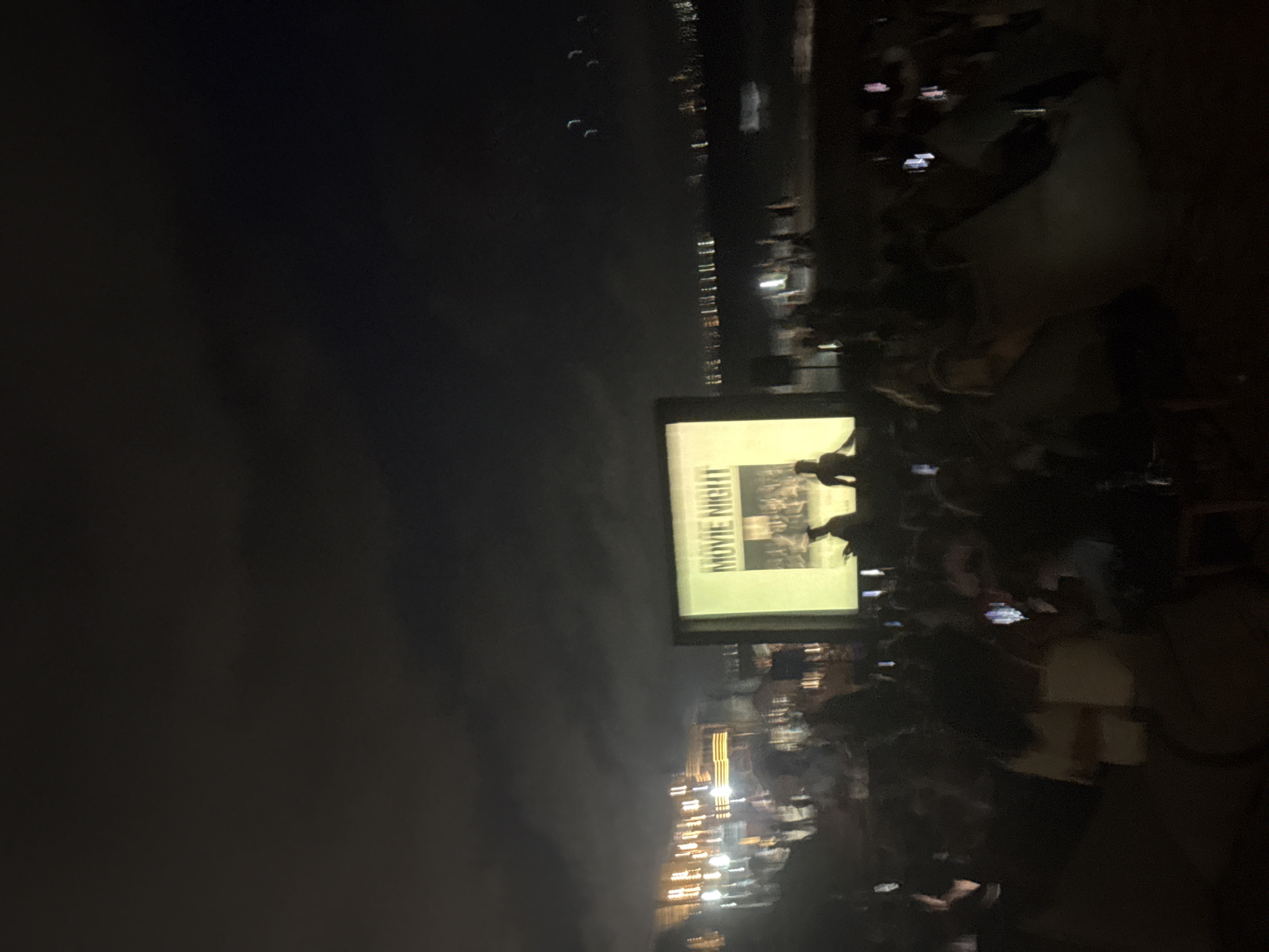 The image depicts an outdoor movie night in an urban setting at night. A large screen displaying "MOVIE NIGHT" is visible, with a crowd of people gathered to watch the film. Many people are holding up their phones, possibly to record the event. In the background, blurred city lights suggest a waterfront location in the city of Unknown. The overall scene conveys a community gathering and a relaxed atmosphere.