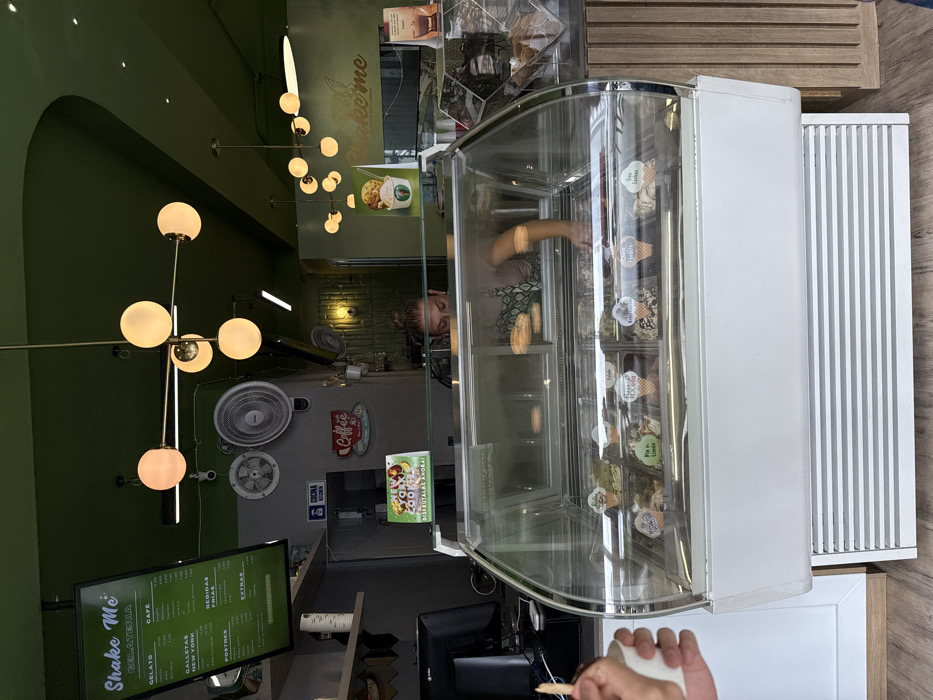 The image depicts the interior of "Shake Me Gelateria," a dessert shop located in AMCO, Area Metropolitana Centro Occidente, Colombia. The scene is characterized by a vibrant green ceiling and upper walls, complemented by light wood paneling and flooring.
In the foreground, a curved glass display freezer showcases various gelato flavors, each labeled with a small cone-shaped marker. To the left, a person's hand holds a white cup with a wooden spoon, suggesting a recently served or purchased item.
Behind the counter and the freezer, a female employee is partially visible, wearing a light-colored shirt and a green patterned apron. She is actively reaching into an area behind the main display, possibly preparing an order or attending to items. Above the counter, a black monitor for a point-of-sale system is visible.
The environment includes two modern Sputnik-style pendant light fixtures with multiple spherical globes illuminating the space. A large digital menu board on the upper left displays "Shake Me Gelateria" and various categories such as "Gelato," "Cafe," "Postres," "Bebidas Frias," and "Extras," with prices listed in Colombian Pesos. Two wall-mounted fans are visible on the grey wall section. Signage includes "COCINA / KITCHEN," a red "Coffee" sign, and a "New York Cookies / DISFRÚTALAS AHORA!" promotional sign. A promotional image for a dessert cup is also displayed to the right of the main counter area.