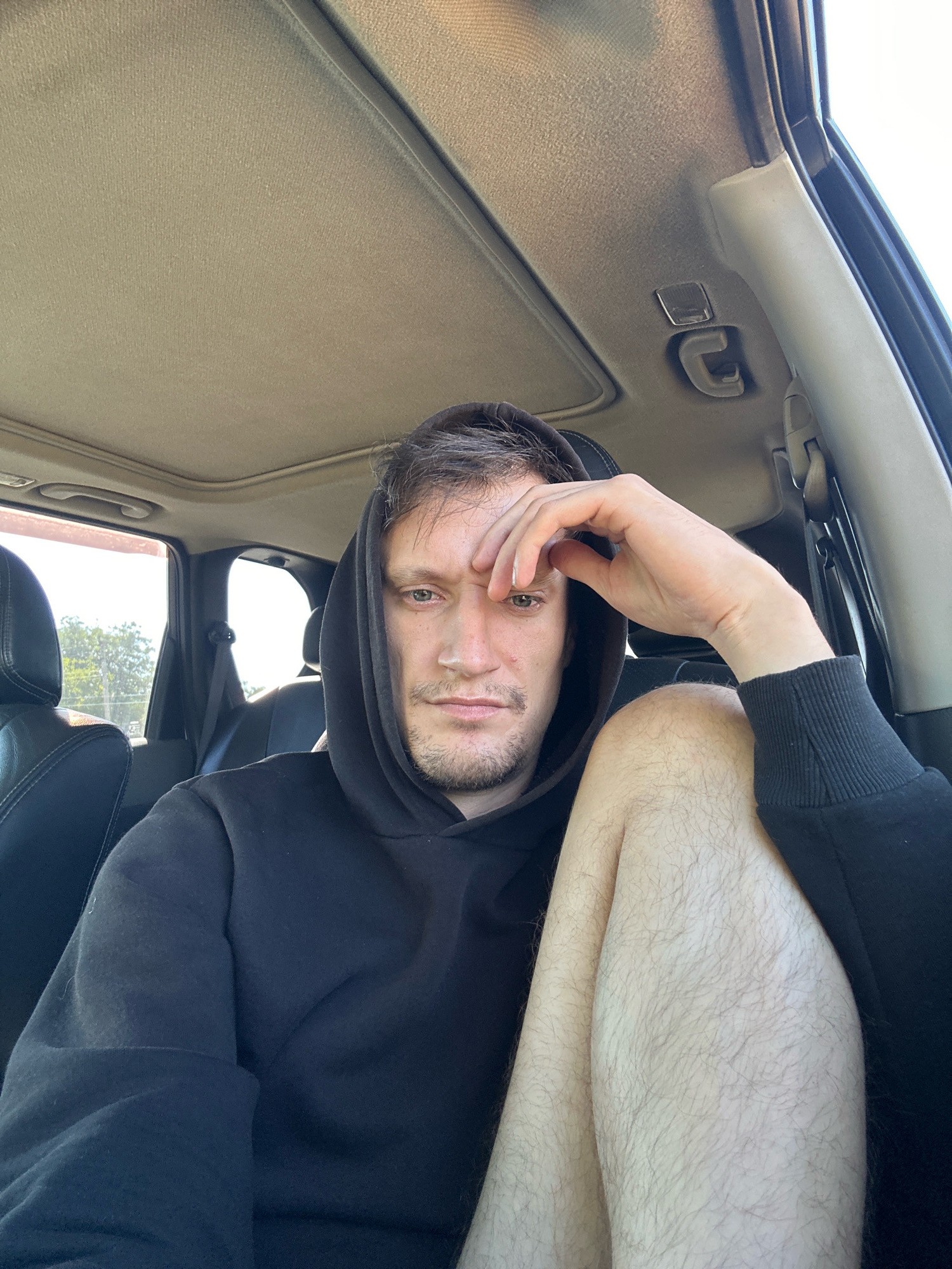 A man sits in the passenger seat of a car, facing the camera. He is wearing a black hooded sweatshirt and has his right hand on his forehead. His left leg is bent and crosses in front of his right. The interior of the car, including the headliner, seats, and a grab handle, are visible. Sunlight streams in through the windows, illuminating the scene. The background outside the windows shows trees and a bright sky, suggesting an outdoor location, possibly in the city of Makiv, Ukraine.