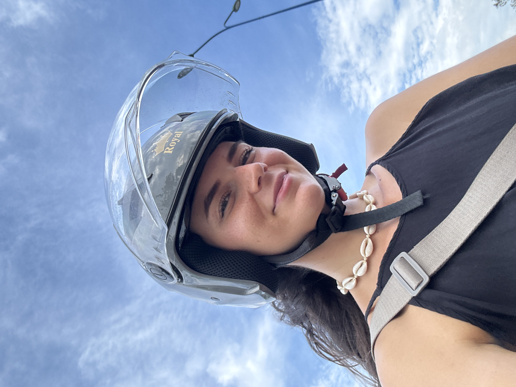 A candid-style, low-angle portrait features a young woman smiling slightly, looking up towards the camera. She is visible from the chest up, wearing a black tank top and a light-colored strap of a bag or purse across her shoulder. Around her neck is a necklace made of white cowrie shells.
The woman is wearing a grey open-face helmet with a clear visor. The brand name "Royal" is distinctly visible in gold lettering on the side of the helmet. Her dark hair is visible beneath the helmet. She has a pleasant expression and appears relaxed.
The background is a bright blue sky with scattered, wispy white clouds, indicating fair weather during the daytime. A street light pole is faintly visible in the upper right portion of the image. The perspective is taken from below, looking up, which highlights the expansive sky. The overall scene suggests an outdoor setting, likely in Mui Ne, Vietnam, possibly while riding or pausing on a scooter or motorcycle.