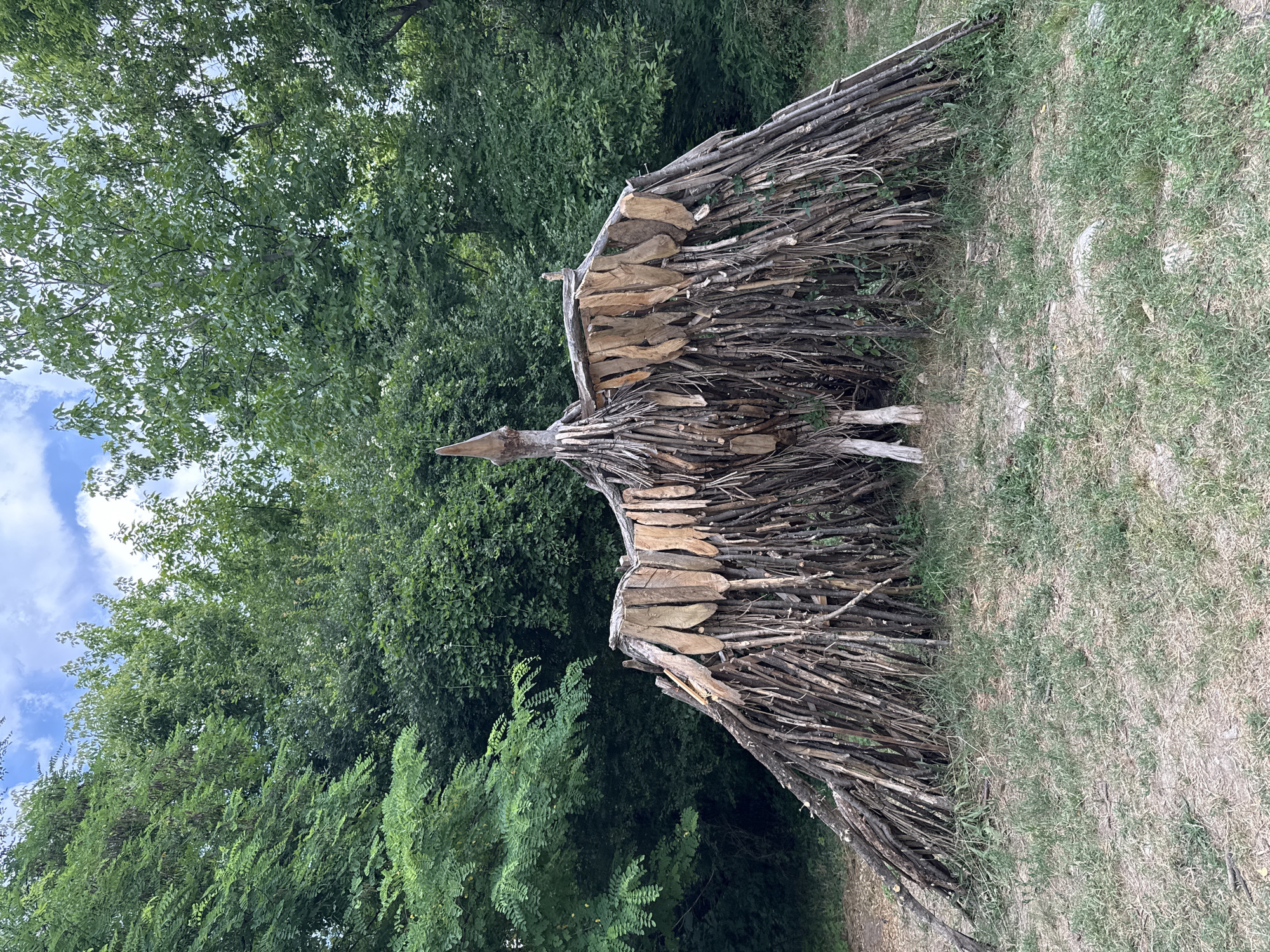 The image depicts an outdoor sculpture of a bird, constructed entirely from natural materials. The sculpture is primarily made of numerous twigs and branches, forming the body, legs, and wings. The wings are adorned with flat, wooden pieces that resemble feathers. The bird sculpture has a pointed beak and stands on two distinct leg structures. It is situated in a grassy area with sparse vegetation and is surrounded by dense green trees, suggesting a natural or park-like setting. The sky above is partially cloudy. This sculpture is located in the park of the Museum of Aviation in Stara Zagora, Bulgaria.