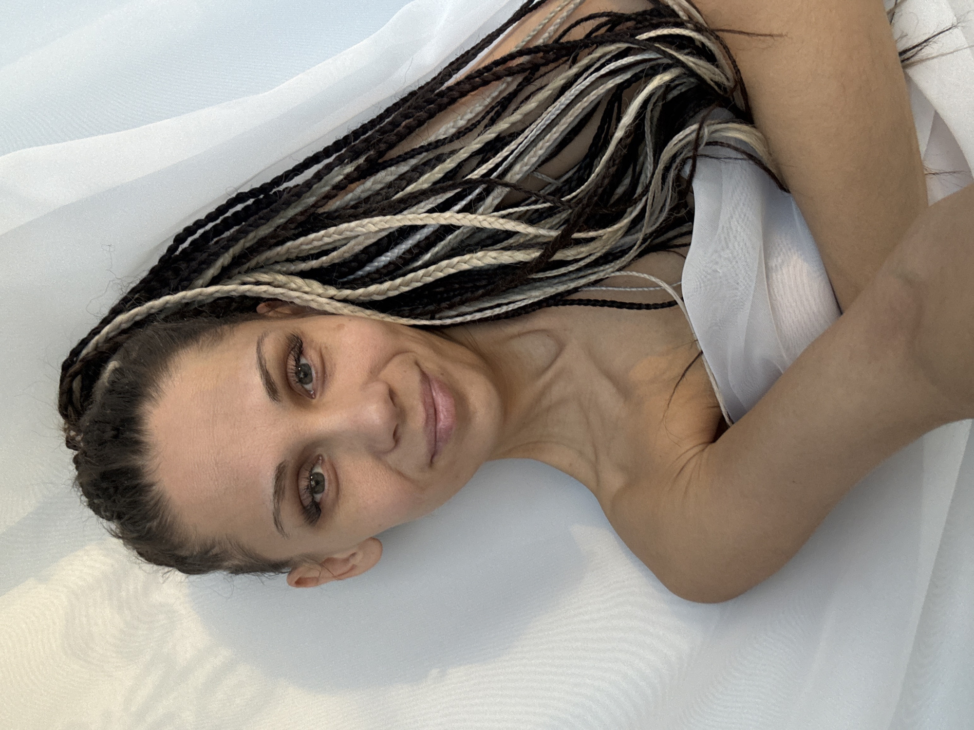 A woman with black and blonde box braids lies on a white fabric background. She is facing the camera with a slight smile. Her left arm is bent, and she is partially covered with white fabric.
There is no information available about the location in the city of Rusanovo, Russia.