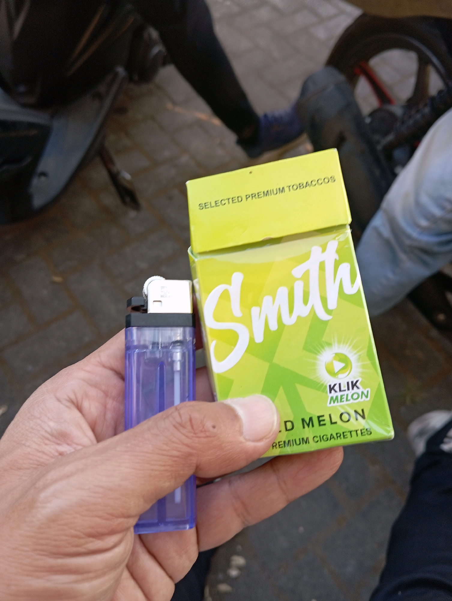 In Sumberrejo, Indonesia, a hand holds a purple plastic lighter and a pack of "Smith" brand cigarettes, "Mild Melon" flavor, with "Selected Premium Tobaccos" printed on the top flap. The cigarette pack is predominantly lime green with white lettering and a circular graphic that reads "KLIK MELON". The background shows a dark-colored motorcycle and a paved walkway.