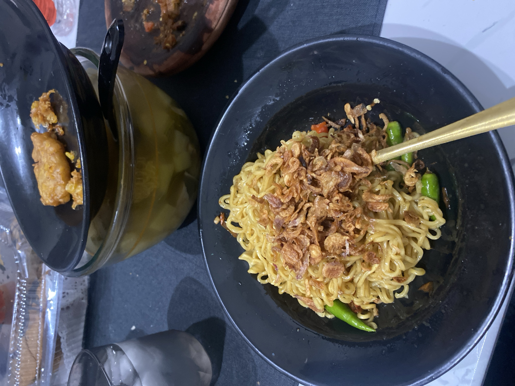 A close-up, eye-level, full shot captures a bowl of instant noodles, topped with fried shallots and garnished with green chilies, positioned on a dark tablecloth. A golden fork rests inside the bowl. In the background, a glass filled with ice water is visible, along with a clear plastic container holding a cake and a glass bowl containing a yellow soup with pieces of food. A black plate with fried food sits atop the soup bowl, and a wooden mortar with chili residue is to the right. The scene appears to be a casual dining setting, likely within a residential or small eatery context in Cibinong, Indonesia, given the presence of common Indonesian food items. The lighting suggests it may be nighttime or an indoor setting with artificial light.