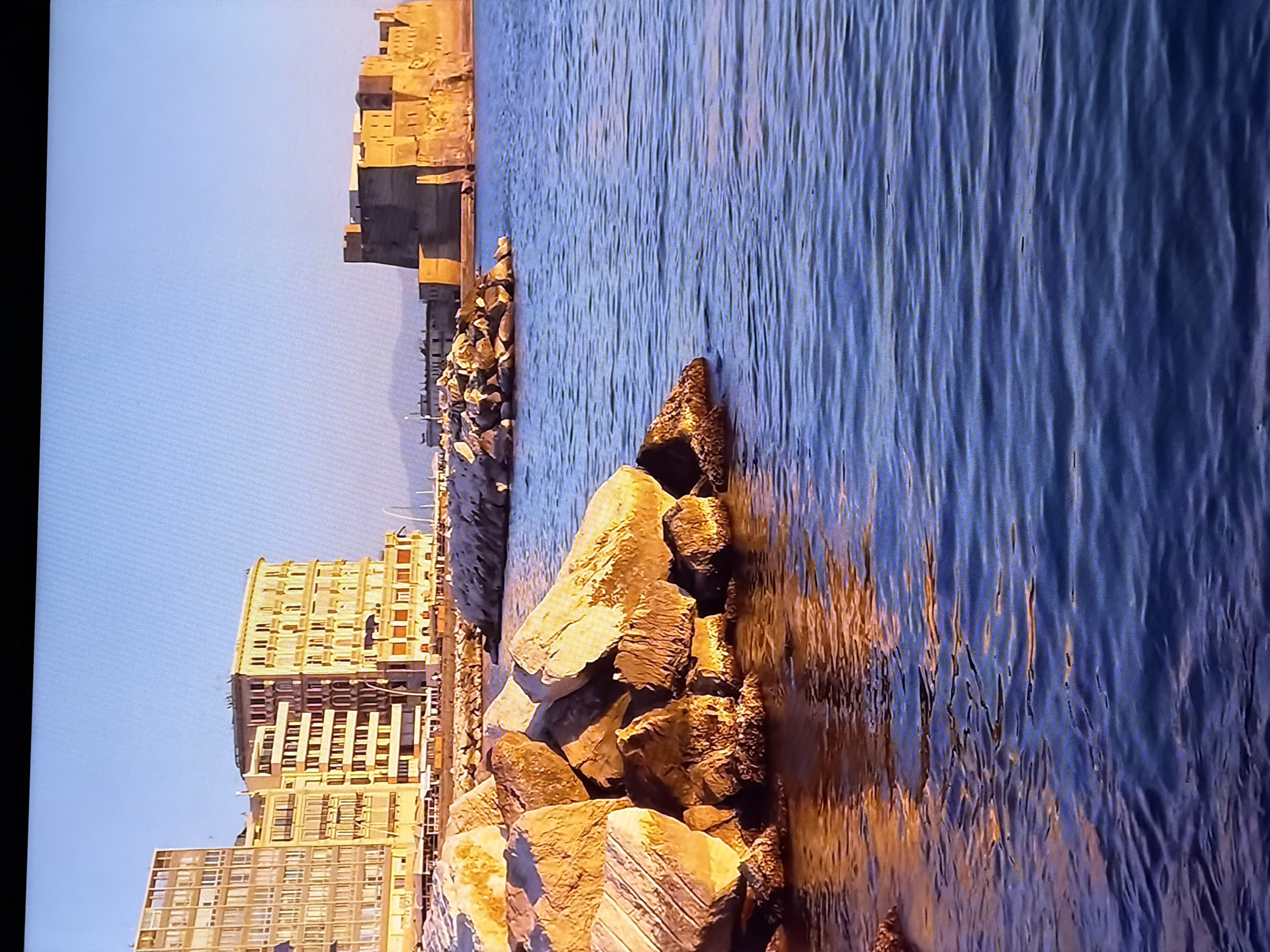 The image shows a coastal scene with buildings and a castle on the horizon, set against a clear blue sky. In the foreground, large rocks are partially submerged in calm blue water, reflecting the warm light of what appears to be sunrise or sunset. The buildings in the background are light-colored and have a layered architectural style. To the right, a large, ancient-looking castle or fortress stands prominently on a rocky outcrop overlooking the sea. The overall impression is one of a scenic, historical waterfront location. The location is not Chirchik, Uzbekistan.