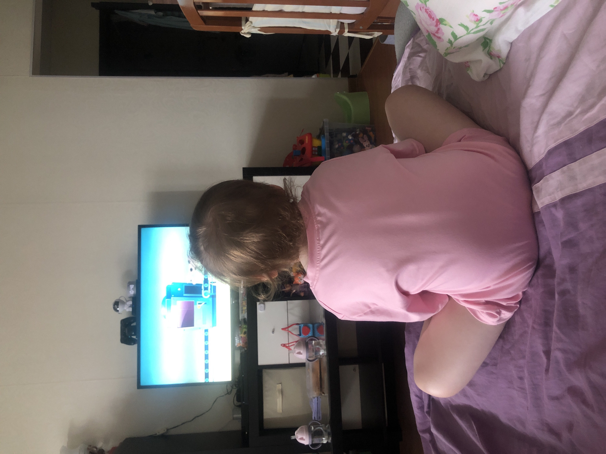 The media file shows a close-up, rotated view of a young child lying on a bed. The child is wearing a pink t-shirt and has light brown hair. The child's legs are bent and crossed, with one leg visible from the knee down. The child is facing away from the camera, towards a television screen that is displaying a colorful, abstract image. The television is mounted on a black shelf unit, which also holds various toys and containers, including a red toy car. The bed is covered with purple and white striped bedding. A pillow with a floral pattern is also visible on the bed. The overall impression is of a child relaxing and watching television in a bedroom. The lighting suggests it is daytime, but there are no strong indicators of weather or specific time of day. No visible text is present in the image.