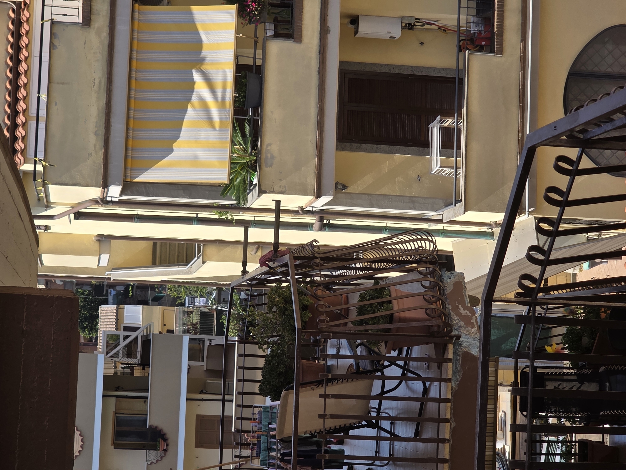The image captures a view of apartment balconies in Ladispoli, Italy. The balconies are constructed of metal railings and are adorned with potted plants and outdoor furniture. On one balcony, a striped awning in yellow and white is extended. Another balcony features a clothes drying rack. The buildings are painted in warm, sun-baked tones of yellow and cream, with terracotta roof tiles visible on the top structures. Sunlight casts shadows across the scene, indicating a bright, clear day. There are no people visible in the image.