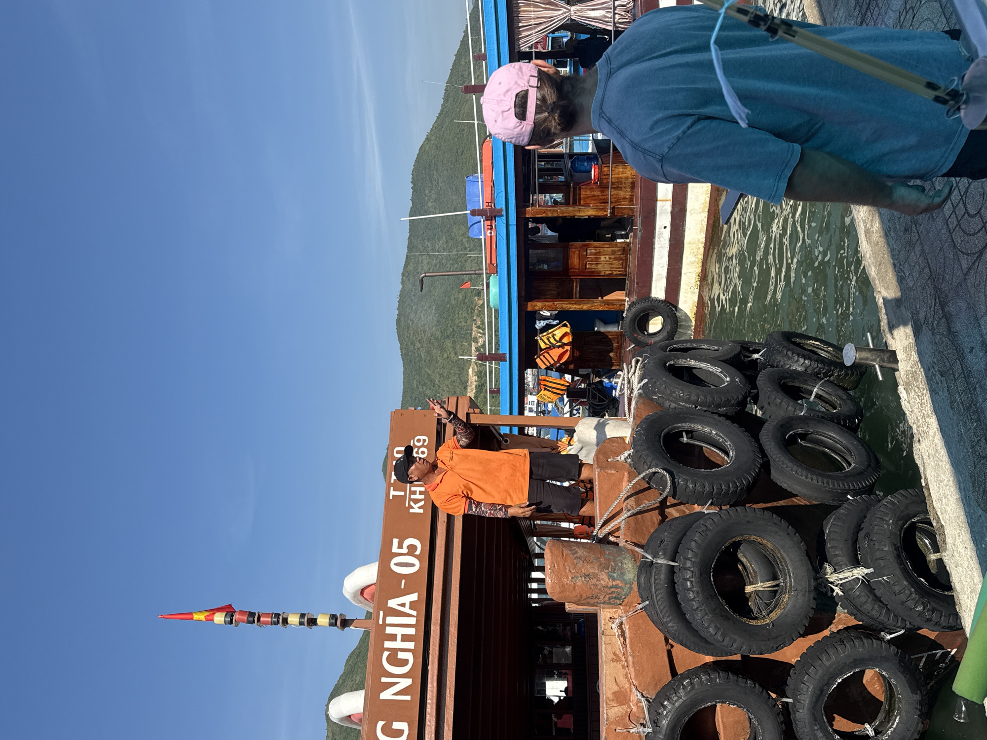 A man in an orange polo shirt and black shorts stands on the deck of a wooden boat in what appears to be a harbor in Nam Nha Trang Ward, Vietnam. He is gesturing with his right hand and appears to be speaking to another person, whose back is to the camera, wearing a blue t-shirt and a pink baseball cap. The boat has the number "05" visible on its side. The water is calm, and there are hills in the background. Several old tires are stacked along the side of the boat, serving as fenders. The sky is clear and blue.