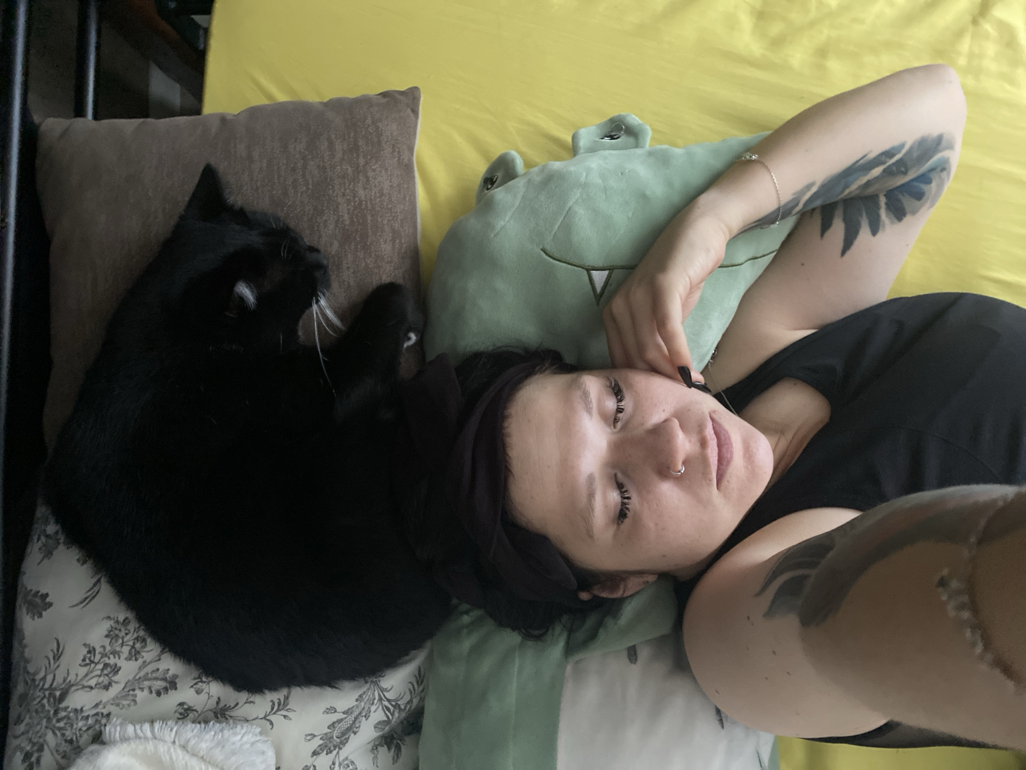 In a bedroom, a black cat rests on top of a woman's head as she lies on a yellow bedspread. The woman is wearing a black tank top and a black headband. She has a nose ring and tattoos on her arms. A green plush toy with a cartoon face is placed under her head. The room is dimly lit, with a patterned duvet and a brown pillow visible. There is no indication that this scene is located in the city of Chaikovsky, Russia.