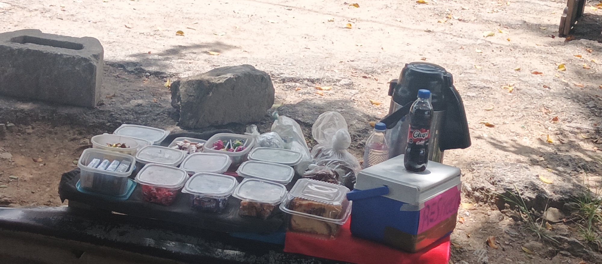 An outdoor vending stall is set up on a dirt and gravel surface, likely in a park or recreational area of Caracas, Venezuela. The stall displays an assortment of food items in clear plastic containers, including what appears to be candies, lollipops, and slices of cake or pastries. A small blue and white cooler sits on the right side of the table, topped with a bottle of "Chupa!" brand soda and a smaller water bottle. Behind the cooler, a large black thermos is visible, suggesting hot or cold beverages are also offered. The background is a dusty, sun-dappled ground with scattered dry leaves and shadows cast from unseen trees. The general atmosphere is that of a simple, casual roadside or park vendor.
