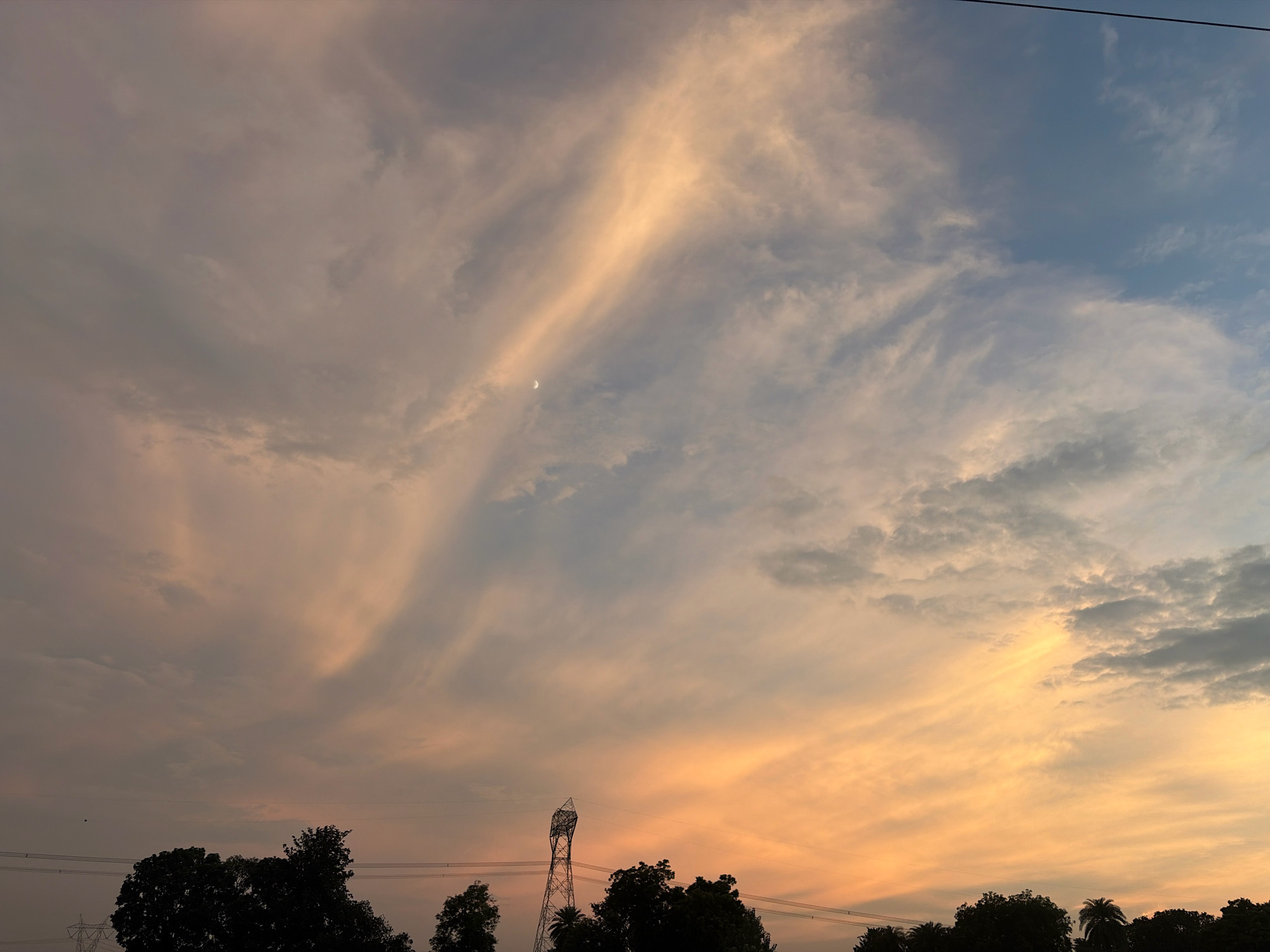 The image depicts a twilight sky over Jhangaha, India. The sky is filled with scattered clouds, tinged with hues of orange, pink, and blue. A sliver of the moon is visible in the upper left quadrant of the frame. Silhouetted against the sky are the tops of trees, power lines, and a tall, lattice-like transmission tower. The overall atmosphere is serene and suggests the transition from day to night.