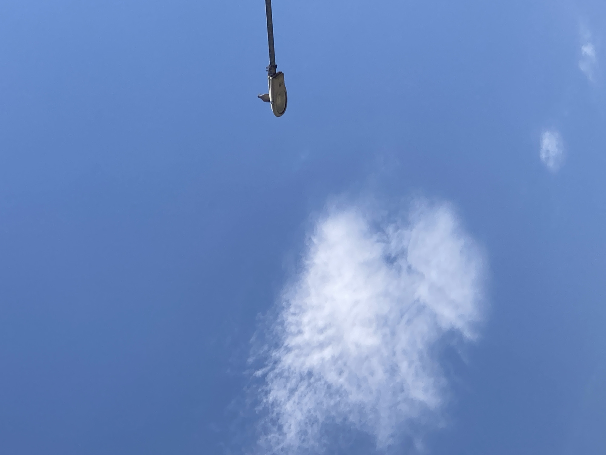 A single bird is perched on a street lamp that extends horizontally across the right side of the frame. The bird is facing left, with its head slightly turned towards the viewer. The street lamp is gray and has a rounded, translucent light fixture. Behind the street lamp and bird, the sky is a clear, bright blue with a few wispy white clouds scattered across the upper left portion of the image. The scene is shot from a low angle, looking up at the sky. The image appears to be taken in an open area, possibly a street or park, with no other objects or structures visible in the frame besides the lamp and sky. This scene could be located in the city of Göreme, Turkey, a region known for its unique geological formations and hot air balloons, which are not visible in this specific photograph.