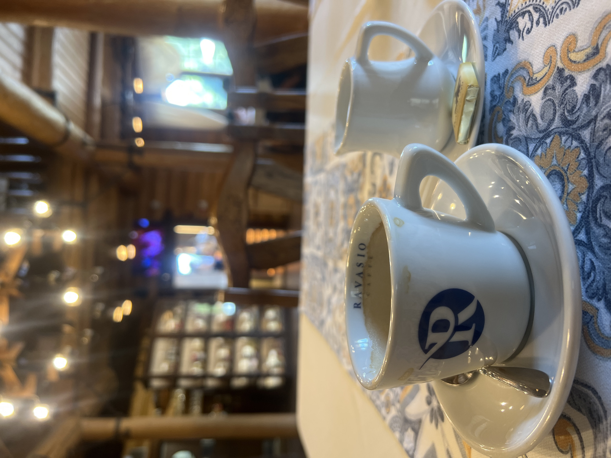 This is a close-up, eye-level shot of a table setting at a cafe in Yaremche, Ukraine. In the foreground, a white coffee cup with a blue "R" logo and the word "RAVASIO" is filled with a dark beverage. A small silver spoon rests on the saucer. To the right, another white ceramic creamer sits on its own saucer, with a small foil-wrapped confection next to it. The table is covered with a white tablecloth adorned with a blue and yellow patterned runner. The background is blurred, showing the interior of a rustic wooden building with exposed beams and lighting fixtures. Shelves filled with items and chairs are also visible.