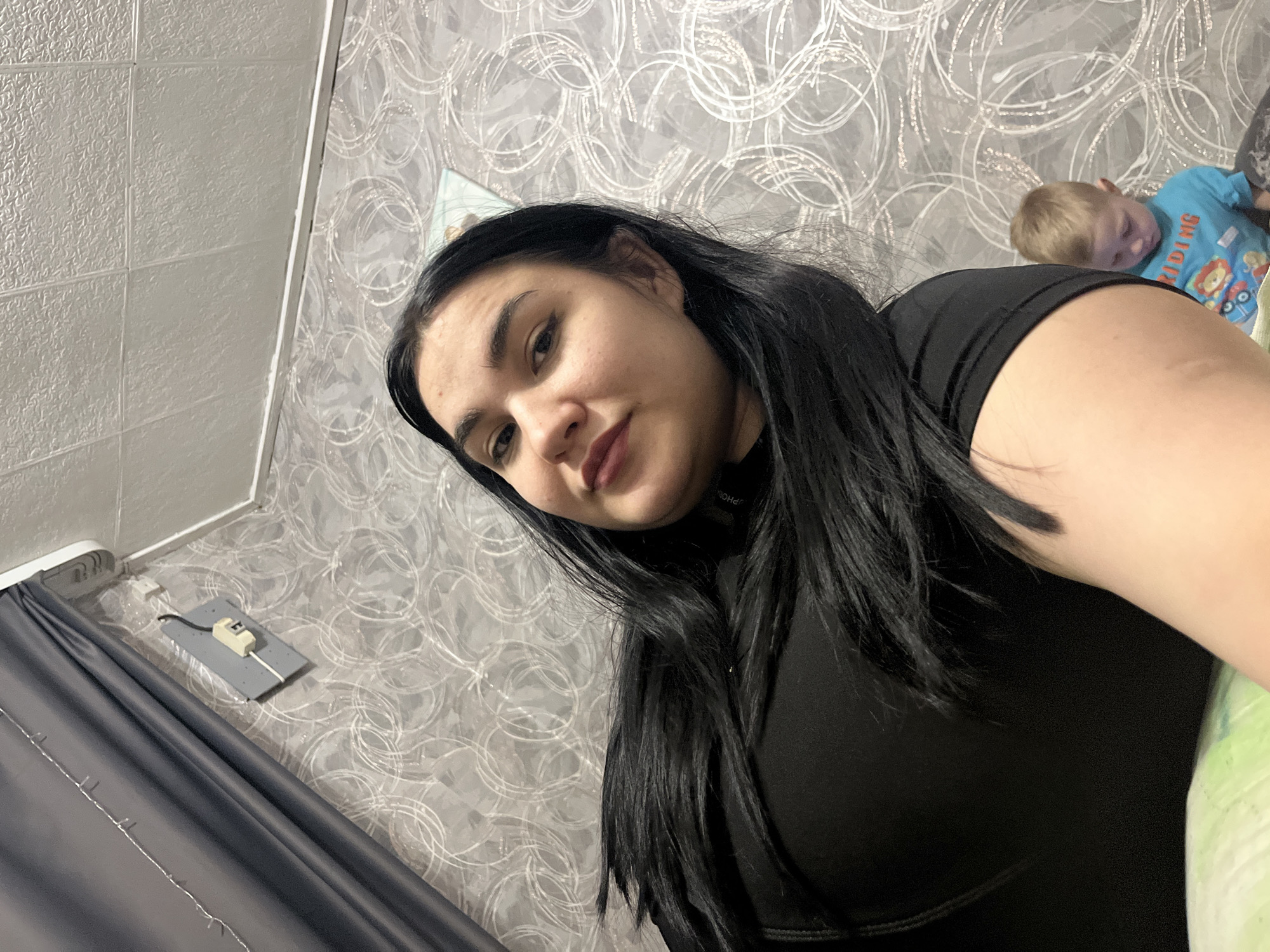This image depicts an indoor scene within a residence located in the city of Chaikovsky, Russia. The primary subject is an adult female, positioned in the foreground and appearing to be captured from a low-angle perspective. She has long, dark hair, is wearing a dark, possibly black, top, and is looking directly towards the camera with a slight head tilt. Her right arm and shoulder are visible in the lower right portion of the frame.
In the mid-ground, to the lower right and partially obscured by the female's arm, a young child with light-colored hair is visible, wearing a light blue t-shirt with a graphic that includes the word "RIDING." The child is looking downwards.
The background consists of a wall covered in gray wallpaper featuring a repeating pattern of white or light-colored swirling circles. To the upper left, a dark gray curtain is present, adorned with a string of small, white fairy lights. Adjacent to the curtain, on the wall, a small, light gray rectangular fixture with a white electrical component and black and white wires is mounted. The ceiling, visible in the upper portion of the image, is white and exhibits a textured, possibly tiled, surface. A segment of light green and white patterned fabric, consistent with bedding or a cushion, is visible in the lower-left foreground. The scene is illuminated by artificial light.