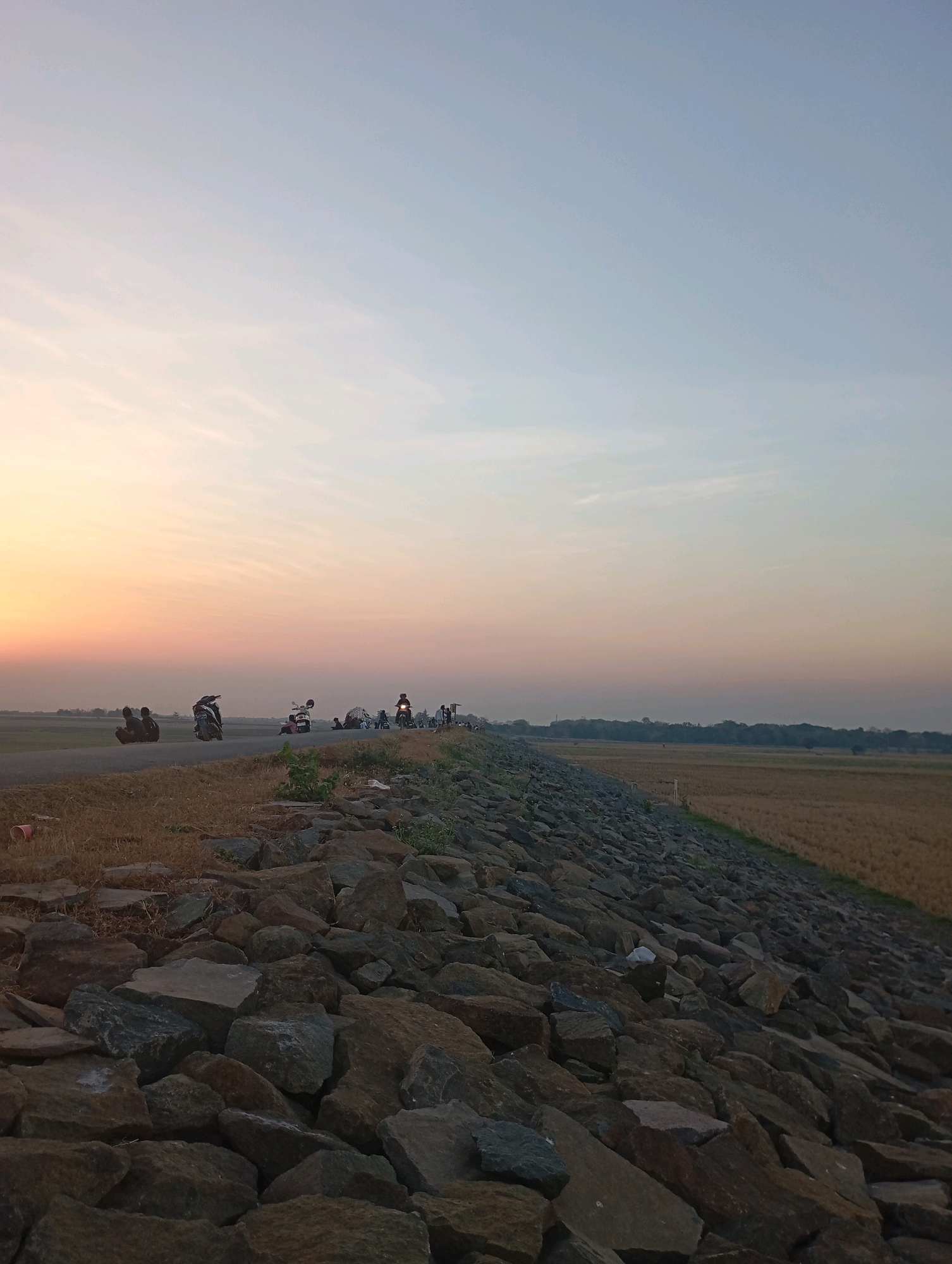 The image depicts an outdoor scene at dusk in Haurkolot, Indonesia. A paved road runs horizontally across the middle ground, bordered on the left by dry grass and on the right by a steep embankment covered in large, irregular stones. Several individuals and motorcycles are present on the road and its immediate surroundings. On the left side of the road, two figures are seated, and a motorcycle is parked. Further along the road, a motorcycle with a rider is visible, as are other motorcycles and individuals gathered in small groups. The sky is a soft gradient of pale orange near the horizon, transitioning to a light blue. Beyond the road and embankment, a field of dry, golden crops stretches into the distance, bordered by a line of trees. The overall lighting suggests either sunrise or sunset, casting a warm glow over the landscape.