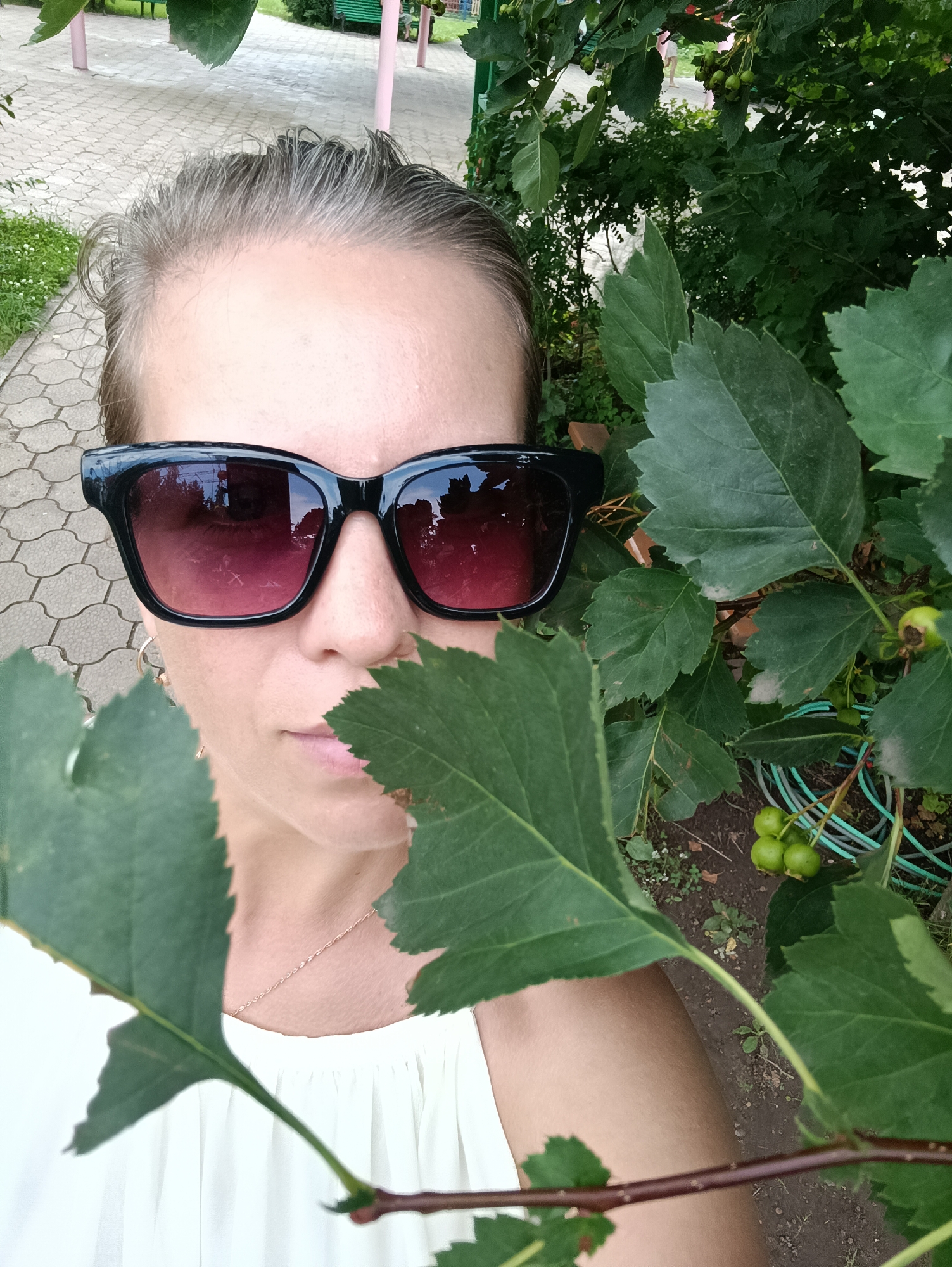 A woman is taking a selfie in an outdoor, urban environment, likely in Chaikovsky, Russia, given the presence of paved walkways and vegetation typical of public spaces. She is partially obscured by the branches and leaves of a tree with small, green fruits. The woman is wearing large, black-rimmed sunglasses with pink-tinted lenses that reflect the surrounding trees and sky. She has her hair pulled back and is wearing a light-colored top. A silver necklace is visible around her neck. The background shows a paved path with a gray, interlocking stone pattern, and further back, hints of park benches and other greenery. The overall lighting suggests it is daytime.