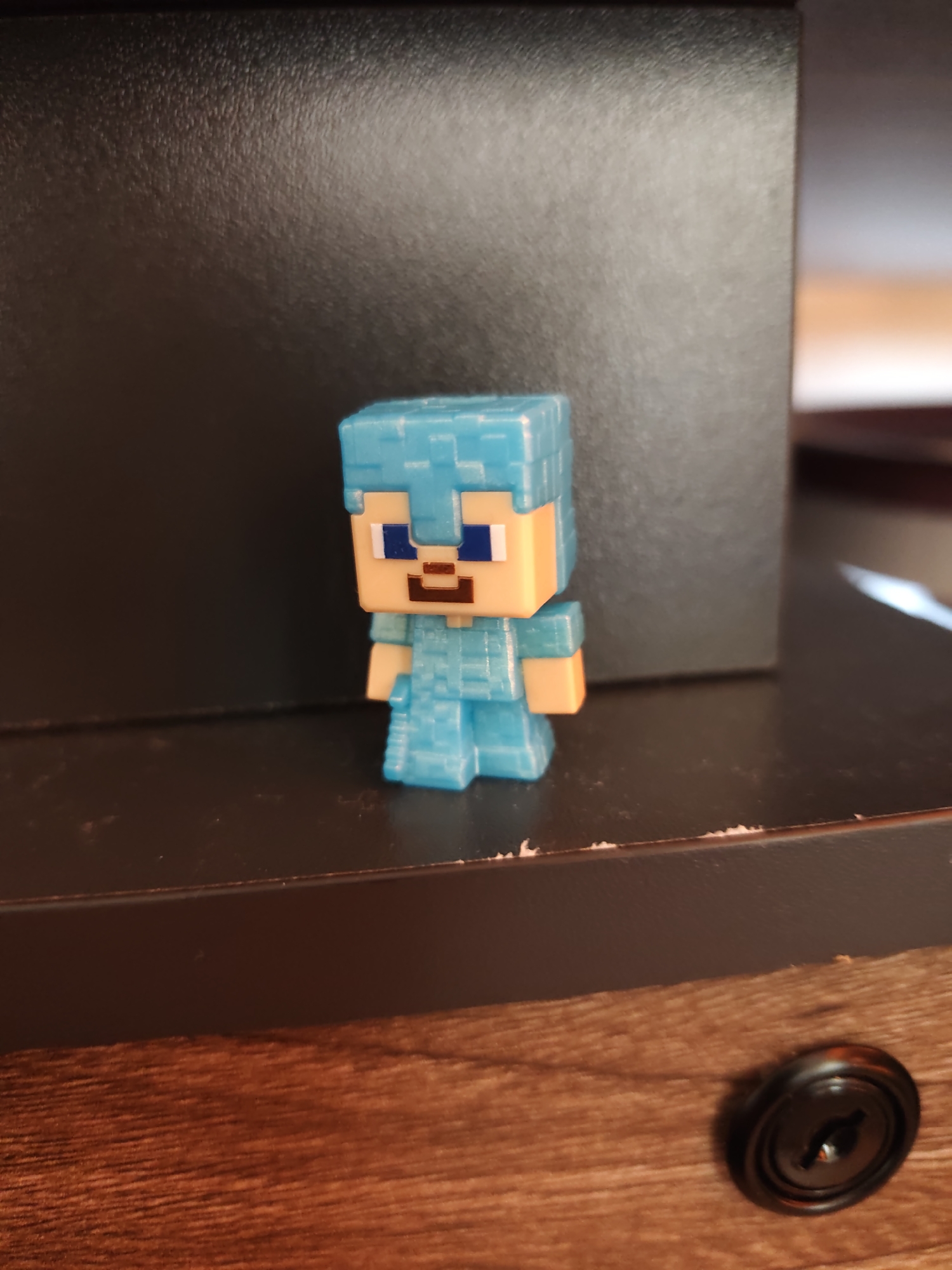 A Minecraft toy figure wearing blue armor is positioned on a dark-colored shelf. The background features a dark, blurry surface. Part of a wooden surface with a black circular knob is visible in the lower right corner. The location in Sofia, Bulgaria cannot be determined from the image.