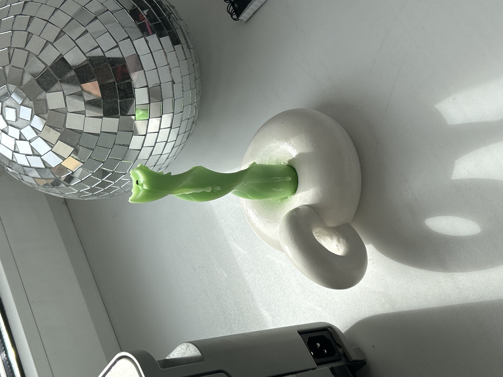 The image shows a disco ball, a white ceramic object resembling a pipe holder with a loop, and a green, twisted candle inserted into the holder. An electronic device with a power input is partially visible in the lower part of the image. The lighting suggests a well-lit indoor environment, possibly a room. The location in Saint Petersburg, Russia is not discernible from the image content.