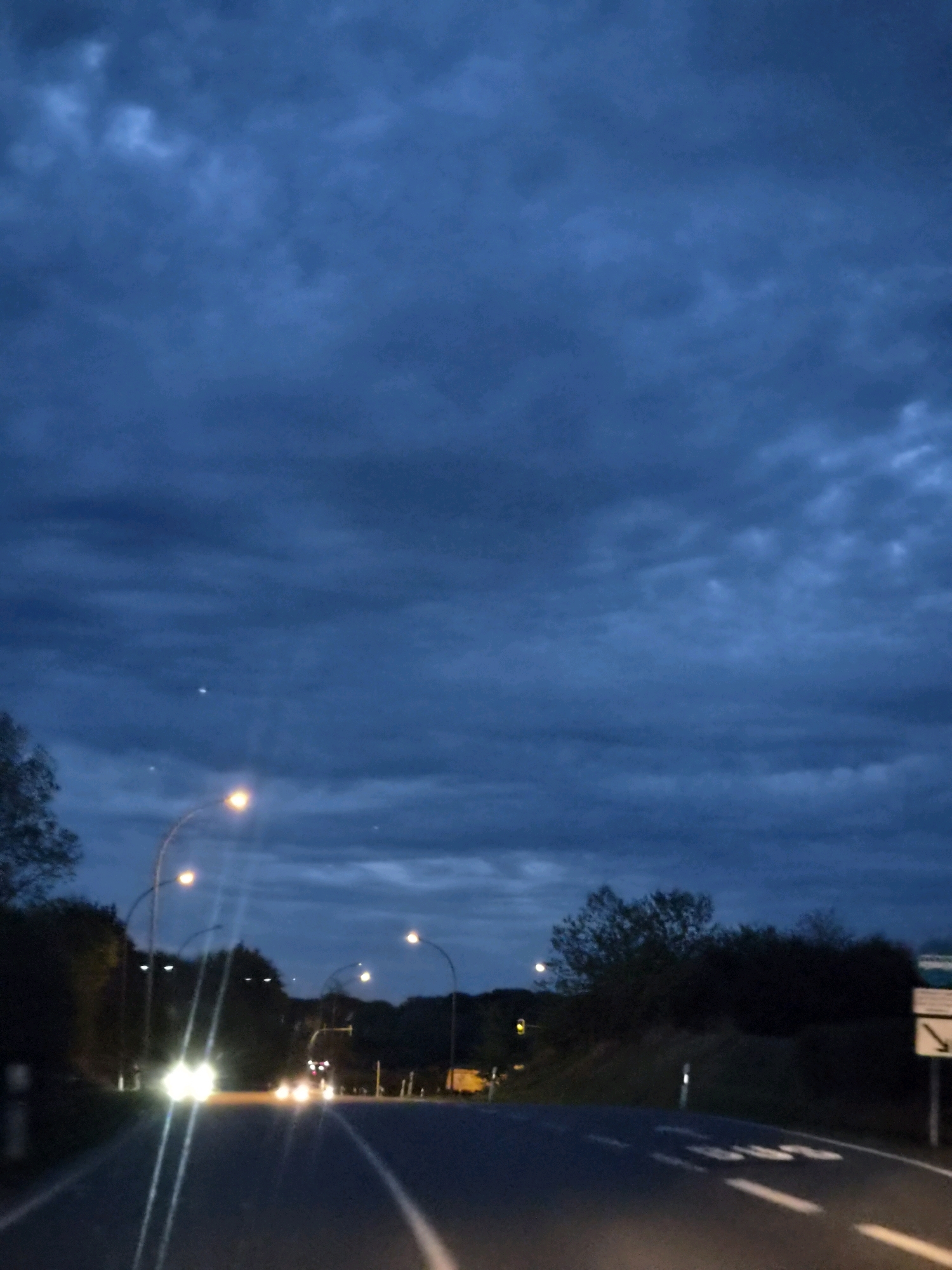 The image depicts a road at dusk or night, with a dark, cloudy sky overhead. Streetlights illuminate the road, and the headlights of approaching vehicles are visible. Trees and vegetation line the sides of the road. The scene suggests a suburban or rural area on the outskirts of a city, given the presence of streetlights and a road that appears to be a main thoroughfare. There are no visible people in the image, and the focus is on the road and the atmospheric conditions. The overall impression is one of a journey or transit through an area as daylight fades.