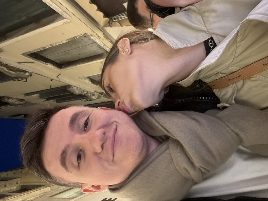 This close-up, low-light image captures two individuals in a narrow street or alley in Venice, Italy, likely at night. The subject on the left is a male, facing forward with a slight smile, looking directly into the camera. He has short brown hair and is wearing a light beige scarf wrapped around his neck, over a dark jacket and a white undershirt. To his right, a female leans her head upwards, puckering her lips towards the male's left cheek without making contact. She has light brown hair, a stud earring in her visible right ear, and is wearing a light tan or beige collared jacket, with a tan leather shoulder bag strap crossing her chest. A third individual is partially visible behind the female on the far right. The background features typical Venetian architecture: tall, light-colored stucco buildings with distinct window frames, some with closed dark shutters, rising steeply on both sides of the narrow passage. An awning, possibly striped, is visible over a ground-floor structure on the right. The scene is illuminated by artificial light, and a dark blue night sky is visible between the building tops.