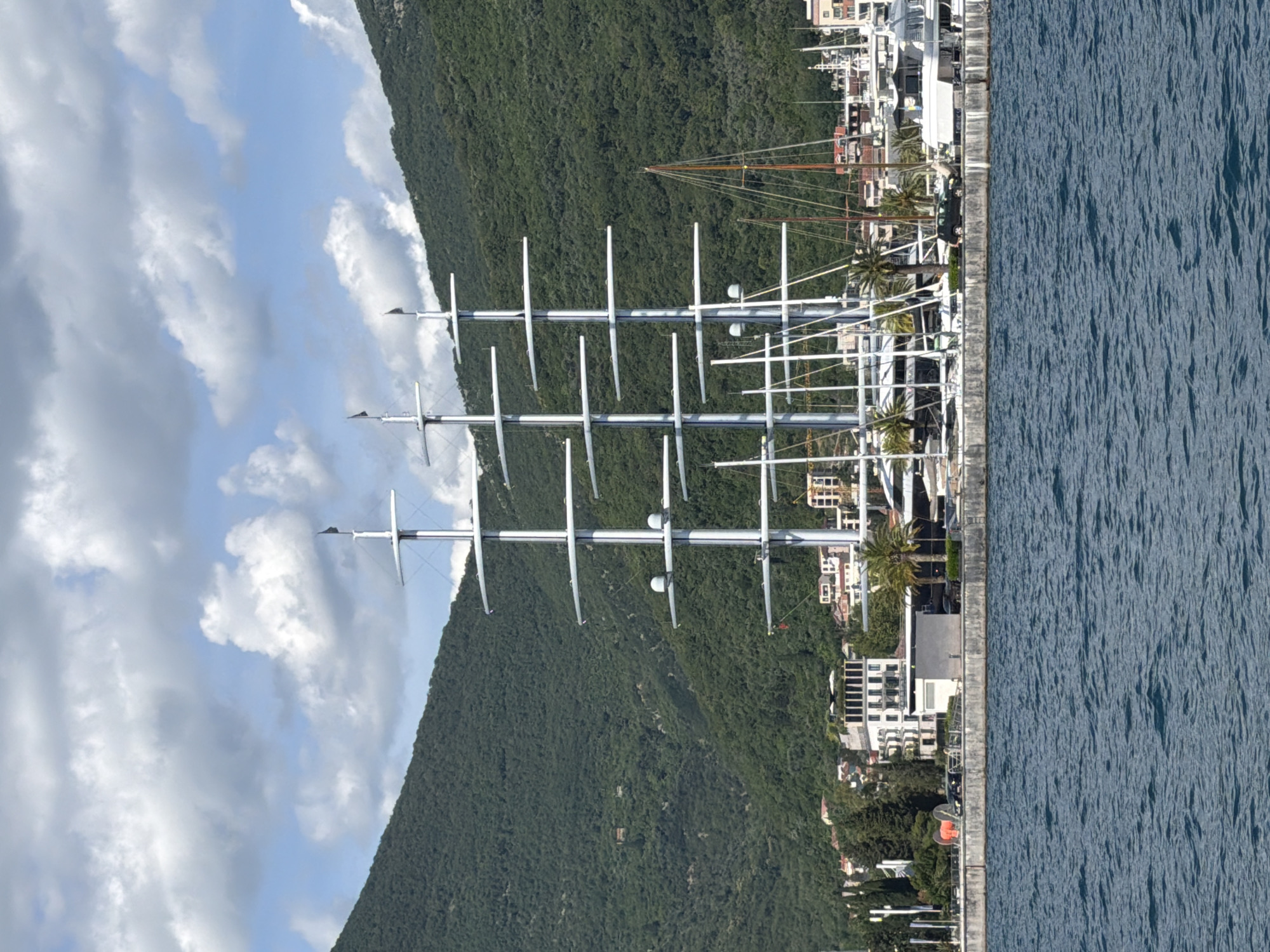 The image depicts a waterfront scene in Tivat, Montenegro. Dominating the foreground is a large, multi-masted sailing vessel with numerous booms extending horizontally from its tall, silver masts. Behind the vessel, a stone embankment separates the water from a promenade area. Along the promenade, buildings of various architectural styles are visible, including modern white structures and older, more traditional buildings. Several palm trees are interspersed among the buildings. Further in the background, lush green hills rise steeply, covered in dense vegetation, with scattered buildings visible on their slopes. The sky above is partly cloudy, with bright white cumulus clouds against a blue sky. The water in the foreground is a deep blue with visible ripples, indicating a slightly choppy sea. Several other smaller boats and yachts are docked near the main vessel.
