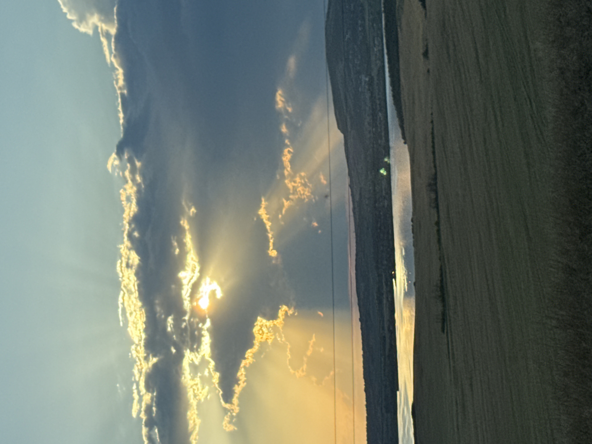 The image depicts a scenic landscape likely taken at dusk, with the sun setting behind dramatic clouds. Sunbeams pierce through the clouds, casting a warm glow on the scene. In the foreground, a field of golden-brown vegetation stretches across the landscape. A body of water, possibly a lake or river, is visible in the mid-ground, reflecting the warm light of the setting sun. Beyond the water, a hill covered in trees and vegetation rises in the distance. Two thin, horizontal wires, likely power lines, are strung across the sky in the mid-ground. The sky above is a gradient of blue and yellow, with the sun partially obscured by dark, textured clouds. The overall impression is one of natural beauty and tranquility. The location is identified as Tvarditsa, Bulgaria.