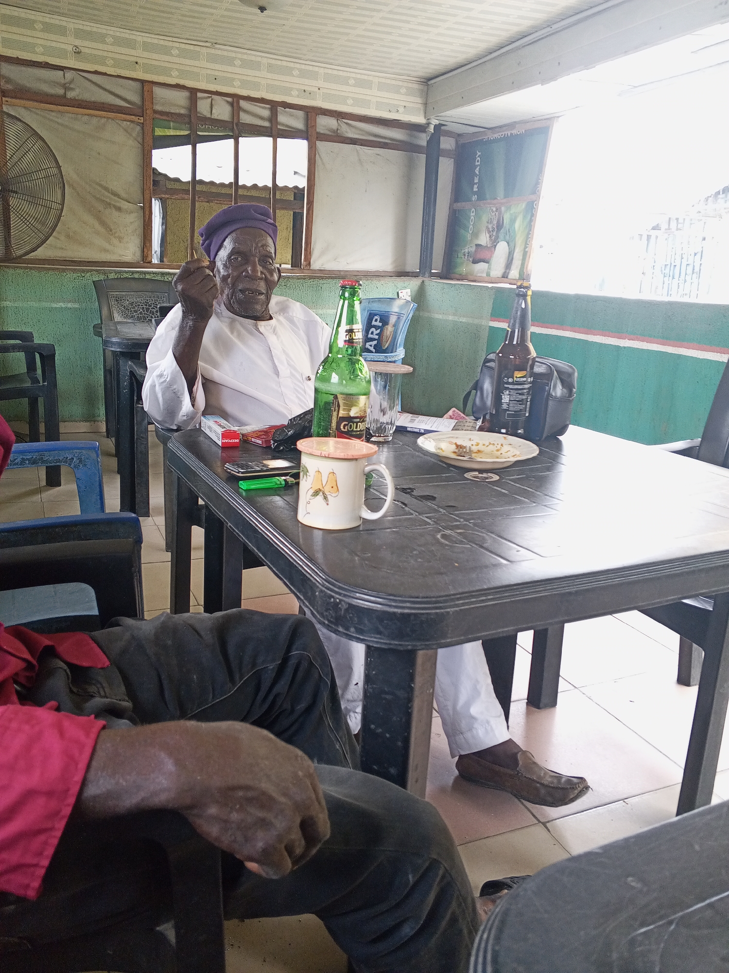 A middle-aged Black man wearing a purple cap and white long-sleeve shirt sits at a black plastic table in an outdoor establishment. He appears to be holding a small object in his right hand. On the table are two bottles of beer, a glass in an ARP ice bucket, a mug with fruit designs, a plate with leftover food, a pack of cigarettes, a lighter, and a cellular phone. The man wears brown loafers. A second person sits in the foreground of the image wearing black pants and a red long-sleeve shirt. The environment consists of green walls with horizontal red and white stripes near the top, a metal window with a white covering, and a ceiling fan on the left side of the frame. A sign on the wall reads "FOOD'S READY." The location is Zango-ndaji, Nigeria.