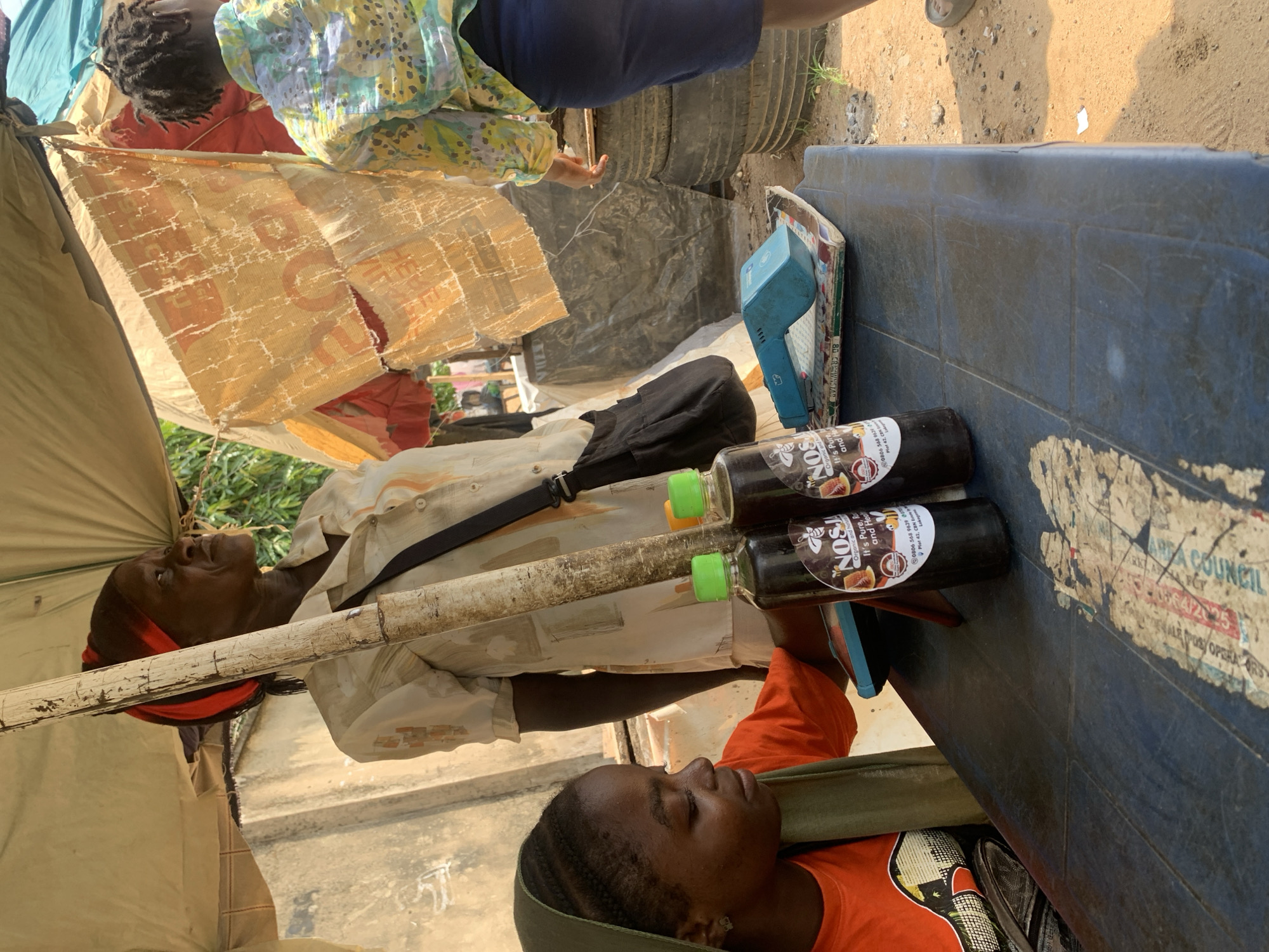 The image depicts an outdoor market stall under a makeshift shelter in Abuja, Nigeria. A woman wearing a red and black headscarf and a cream-colored shirt stands behind a wooden pole. To her left, a young woman wearing an orange t-shirt and a green headscarf is seated. On the table in front of them are two dark glass bottles with green caps, labeled "NOS! Organic and Natural." Next to the bottles is a blue electronic device, possibly a point-of-sale terminal, placed on top of some magazines. In the background, another woman wearing a colorful patterned shirt is partially visible, and stacks of old tires are visible to the right. The ground is dirt and sand. The scene appears to be a typical street-side vendor setup in a bustling urban environment.