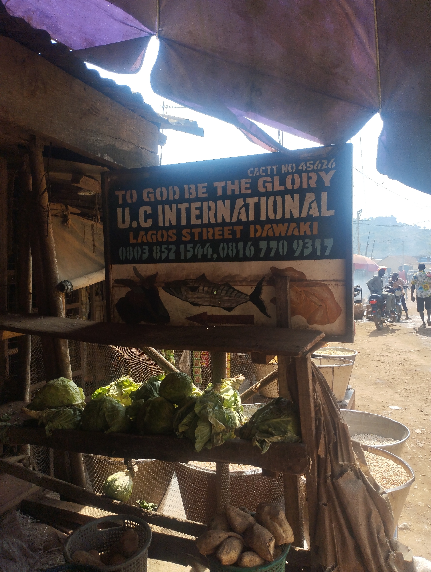 The image captures a small, open-fronted shop, likely in Dutsen Alhaji, Nigeria. A sign displays "TO GOD BE THE GLORY U.C INTERNATIONAL LAGOS STREET DAWAKI" along with two phone numbers. CACTT No. 45626 is at the top right of the sign. Below the text is a drawing of a fish and a piece of meat. The shop's shelves hold cabbages and yam, potatoes in plastic baskets and grains in buckets. Outside the shop, people are walking and one person is on a motorcycle. The buildings are visible in the background. The scene is bathed in sunlight.