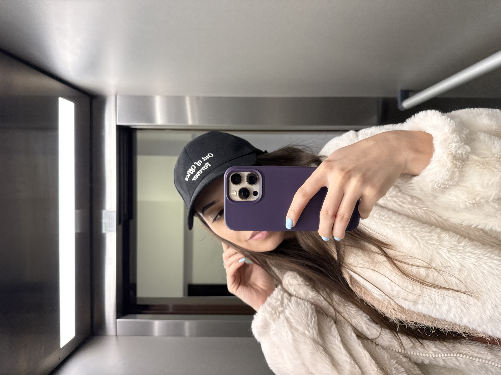 A person is taking a selfie in what appears to be an elevator. The person is wearing a black baseball cap with white writing on it, a light-colored, fluffy jacket, and is holding a purple cell phone. The elevator has brushed metal walls and a light panel on the left. The background visible through the elevator door is out of focus. The location in the city of Plovdiv, Bulgaria, is not evident from the image.