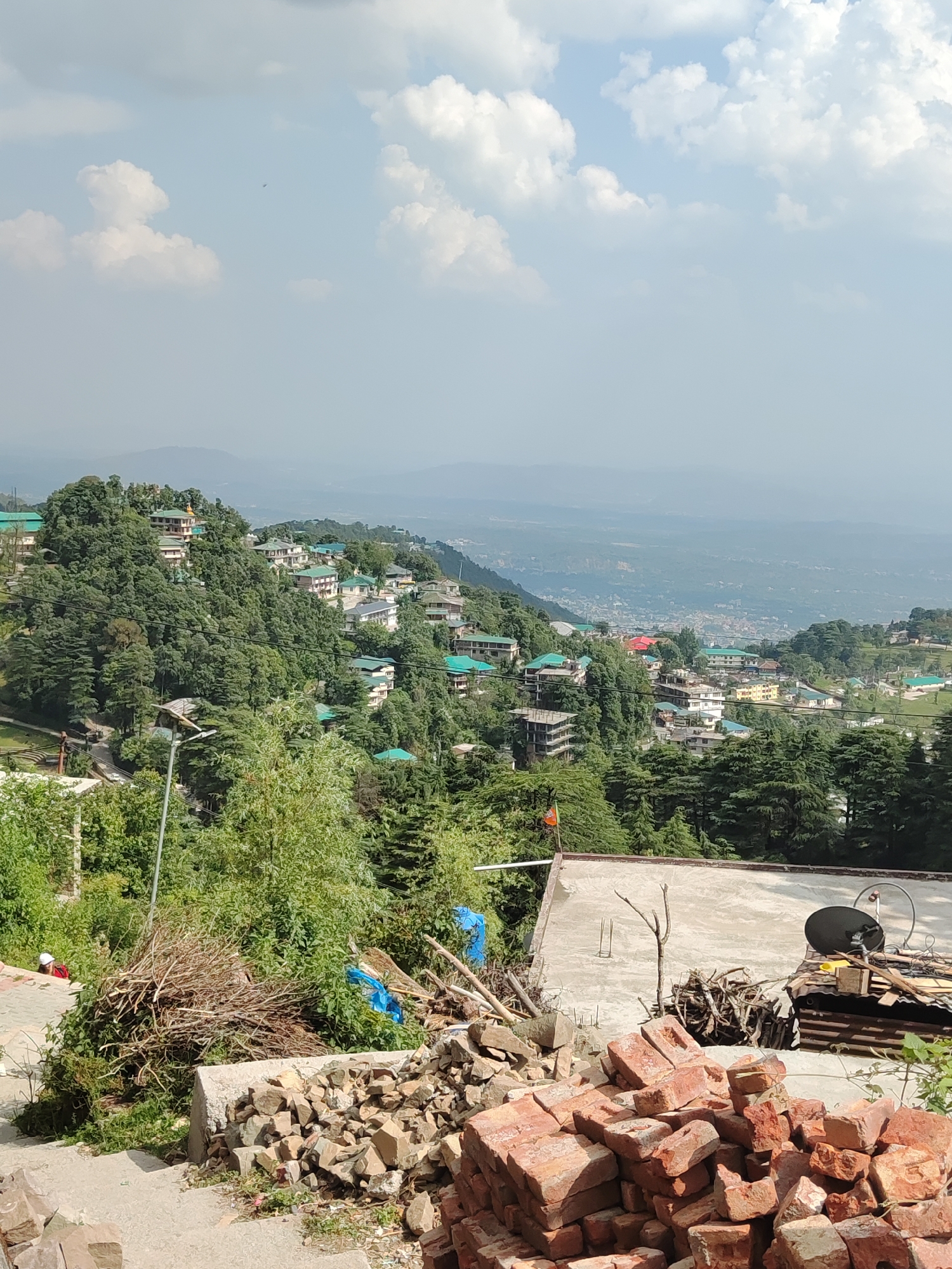 The image depicts a hillside town in Unknown, Unknown, with houses nestled among dense trees. The sky is light blue with scattered white clouds. In the foreground, a pile of red bricks and rocks is visible, along with construction materials and branches. A concrete building with a satellite dish sits behind the debris. Further back, the town sprawls across the hillside, blending into the distant landscape.