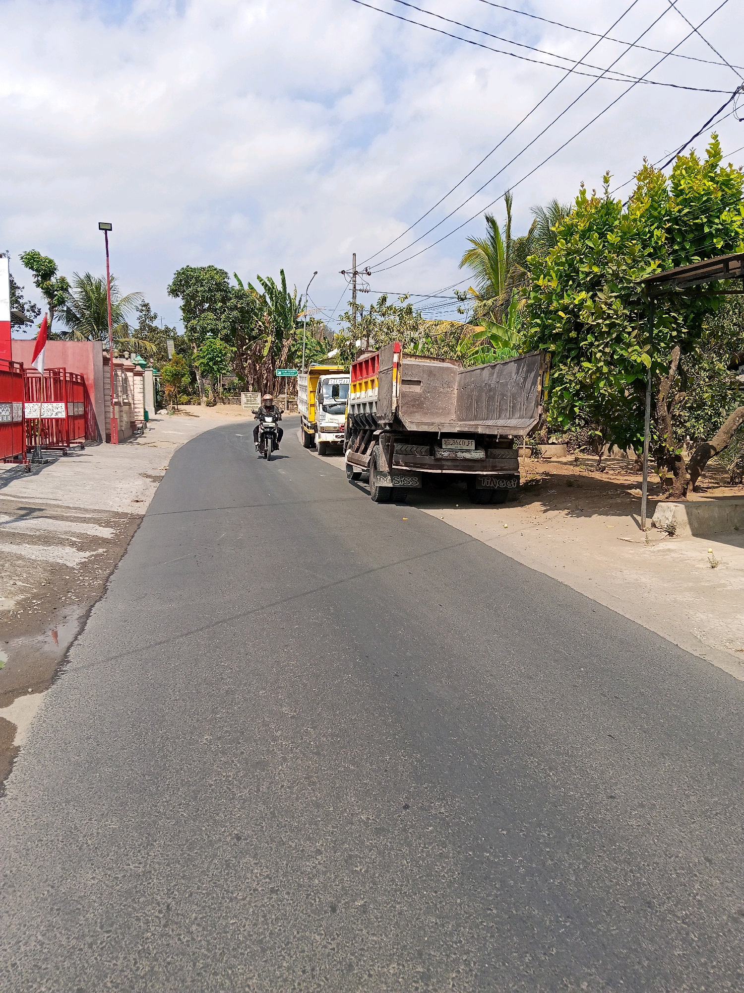 The image depicts a paved road in Bacem, Indonesia, with a motorcycle and two dump trucks present. A person on a motorcycle, wearing a helmet, is riding down the road. To the right of the motorcycle are two dump trucks parked on the side of the road. The dump trucks are grey and yellow. The road is asphalt and appears to be wet in some areas. There are trees and other vegetation on the sides of the road. A red gate with an Indonesian flag is visible on the left side of the image. The sky is partly cloudy. The environment suggests a semi-rural or suburban setting.