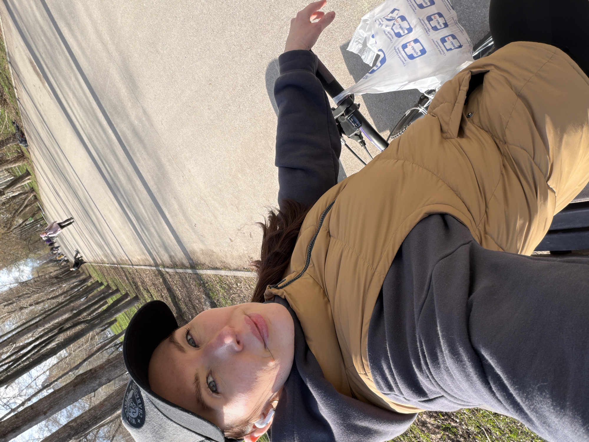 The image is a selfie taken by a woman wearing a gray sweatshirt, a tan puffer vest, and a baseball cap. She is riding a bicycle on a paved path. A plastic bag with the Russian pharmacy brand "apteka.ru" is visible on the handlebars. The path runs alongside a line of trees. In the background, other people can be seen walking along the path. Based on the Cyrillic text on the pharmacy bag, the image was most likely taken in Russia. Further information, such as the specific type of trees, architectural features of the buildings (if any) in the distance, and precise landmarks, would be needed to determine the specific location in Perm, Russia.