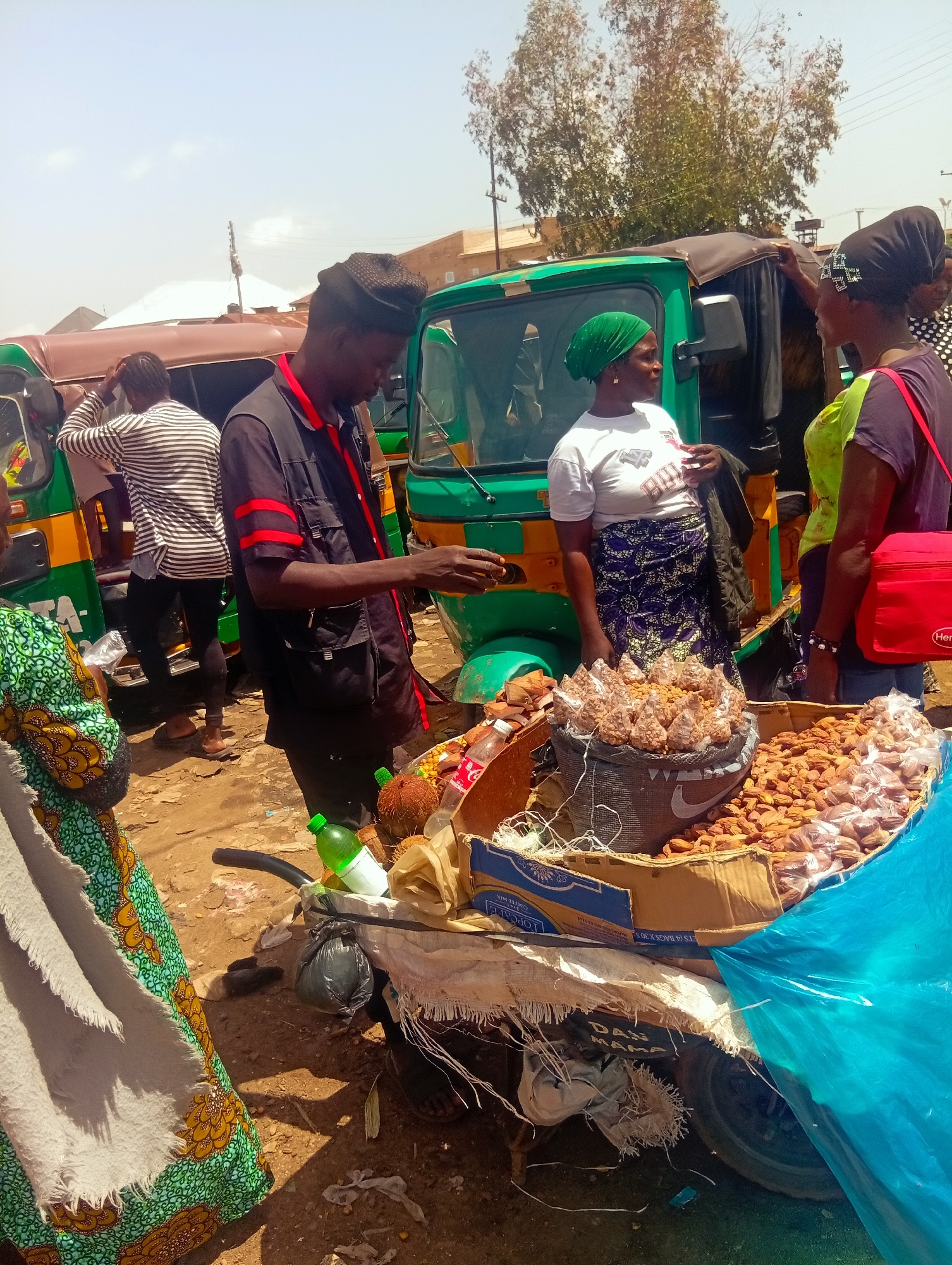 In a busy street market in Bukuru, Nigeria, a vendor, a man wearing a black vest over a red and black striped shirt, stands behind a cart laden with goods. He is interacting with a woman, identified by her green headscarf and white t-shirt. Beside them, two other women in headscarves are engaged in conversation near a green motorized tricycle. The cart displays a variety of food items, including bags of nuts, coconuts, and a bottle of Coca-Cola. The ground is dusty, indicative of a dry, outdoor market environment. The background shows more tricycles and simple structures, suggesting a typical commercial area in a Nigerian city.