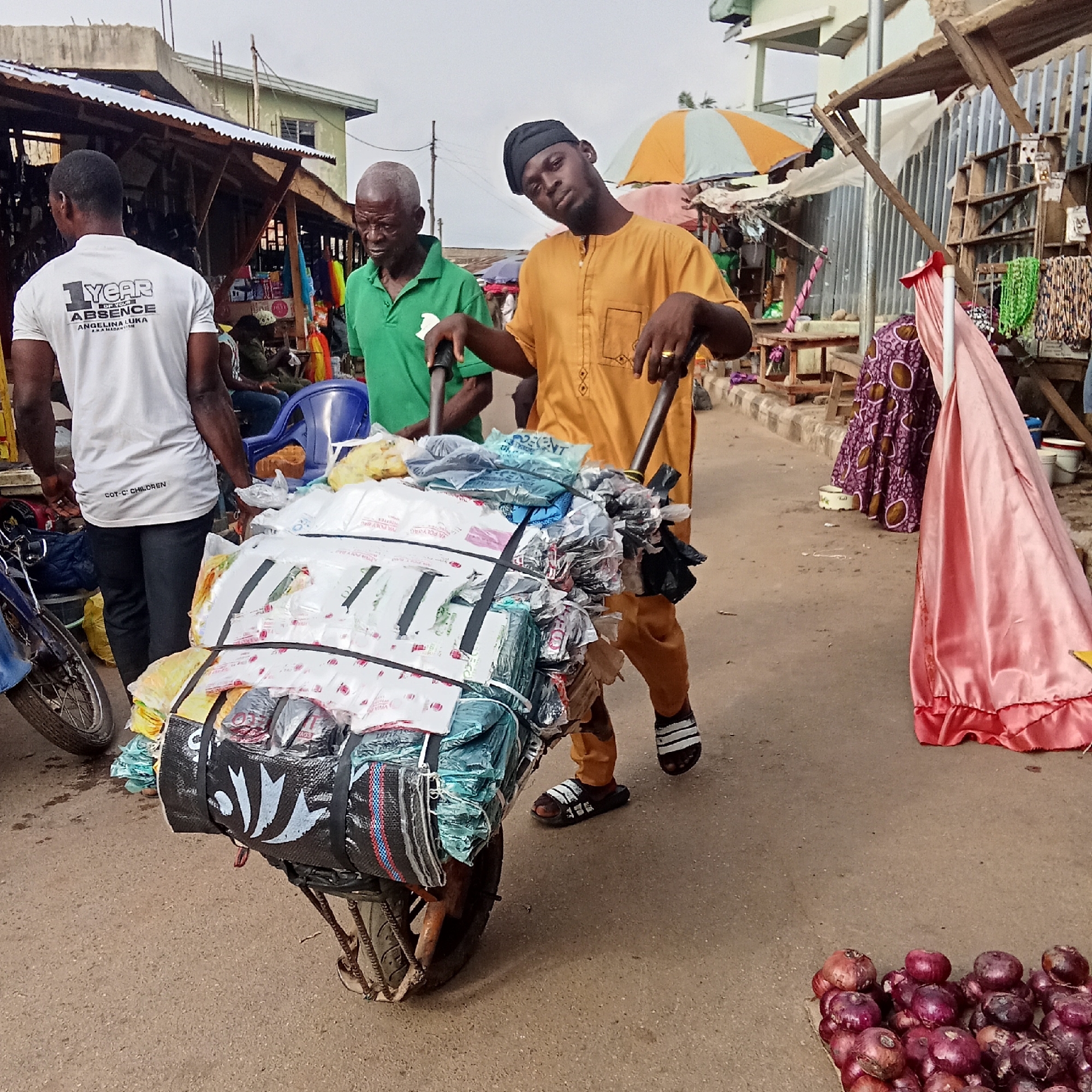 The image captures a street scene in Akwanga, Nigeria. Two men are pushing a heavily loaded wheelbarrow along a paved street. The wheelbarrow is filled with a large assortment of items, many wrapped in plastic bags and secured with straps. A third man is standing to the left.
The man closest to the camera is wearing a mustard-colored tunic-style top and matching pants, a black cap, and sandals. The older man assisting in pushing the wheelbarrow is wearing a green polo shirt. The third man is wearing a white shirt with black text on the back, black pants, and is facing away from the camera.
The background reveals a bustling street market with various stalls displaying goods. A motorcycle is parked next to the first man. To the right of the frame, clothes are hanging from a stall. In the foreground, there is a pile of red onions. The weather appears to be overcast.